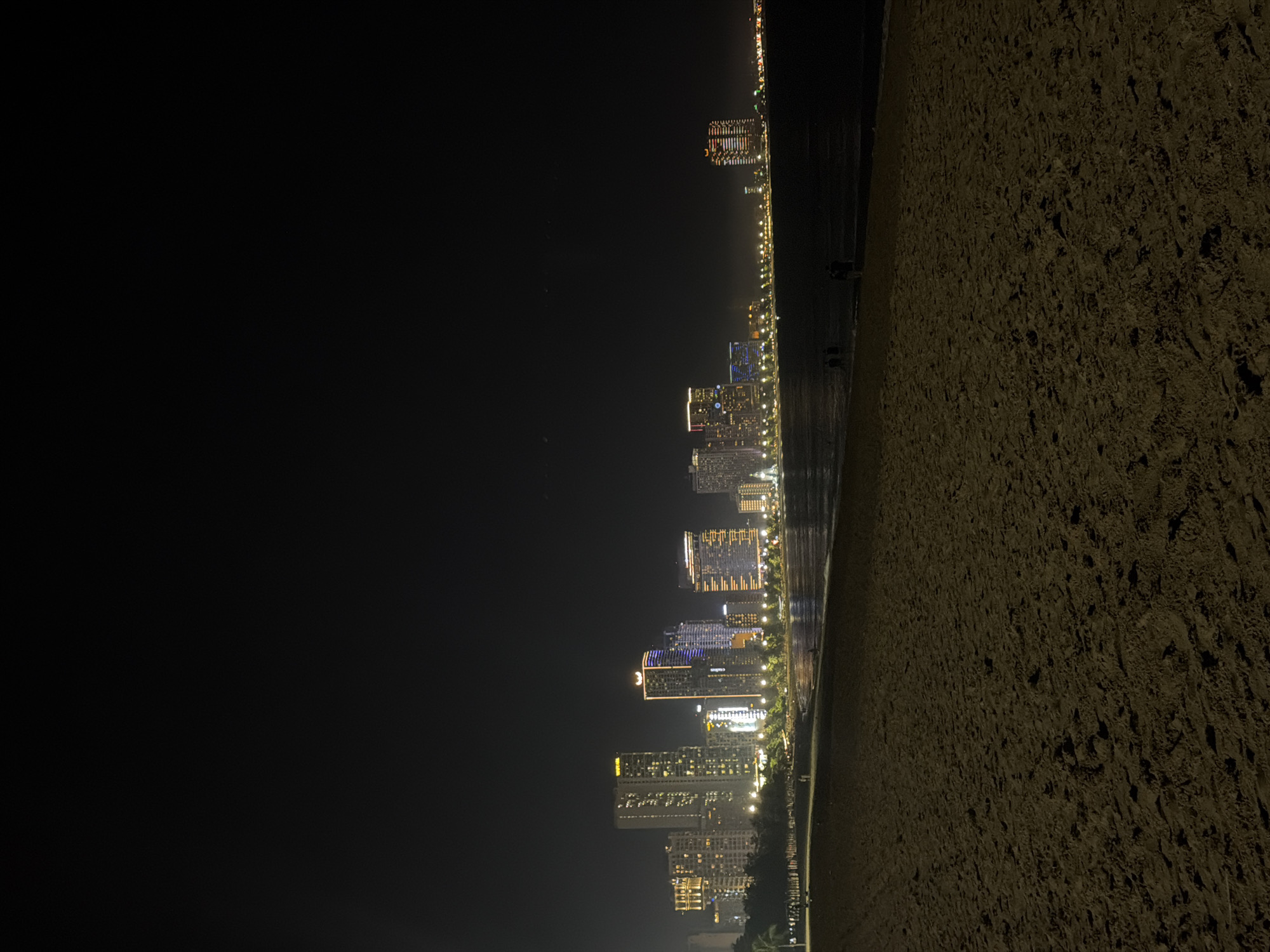 The image depicts a nighttime view of a coastline in Nha Trang Ward, Vietnam. The city skyline is illuminated with numerous lights from buildings along the shore. To the left is a large dark area depicting the nighttime sky. To the right is the beach, which is also darker in tone. The water line is between the beach and the skyline.