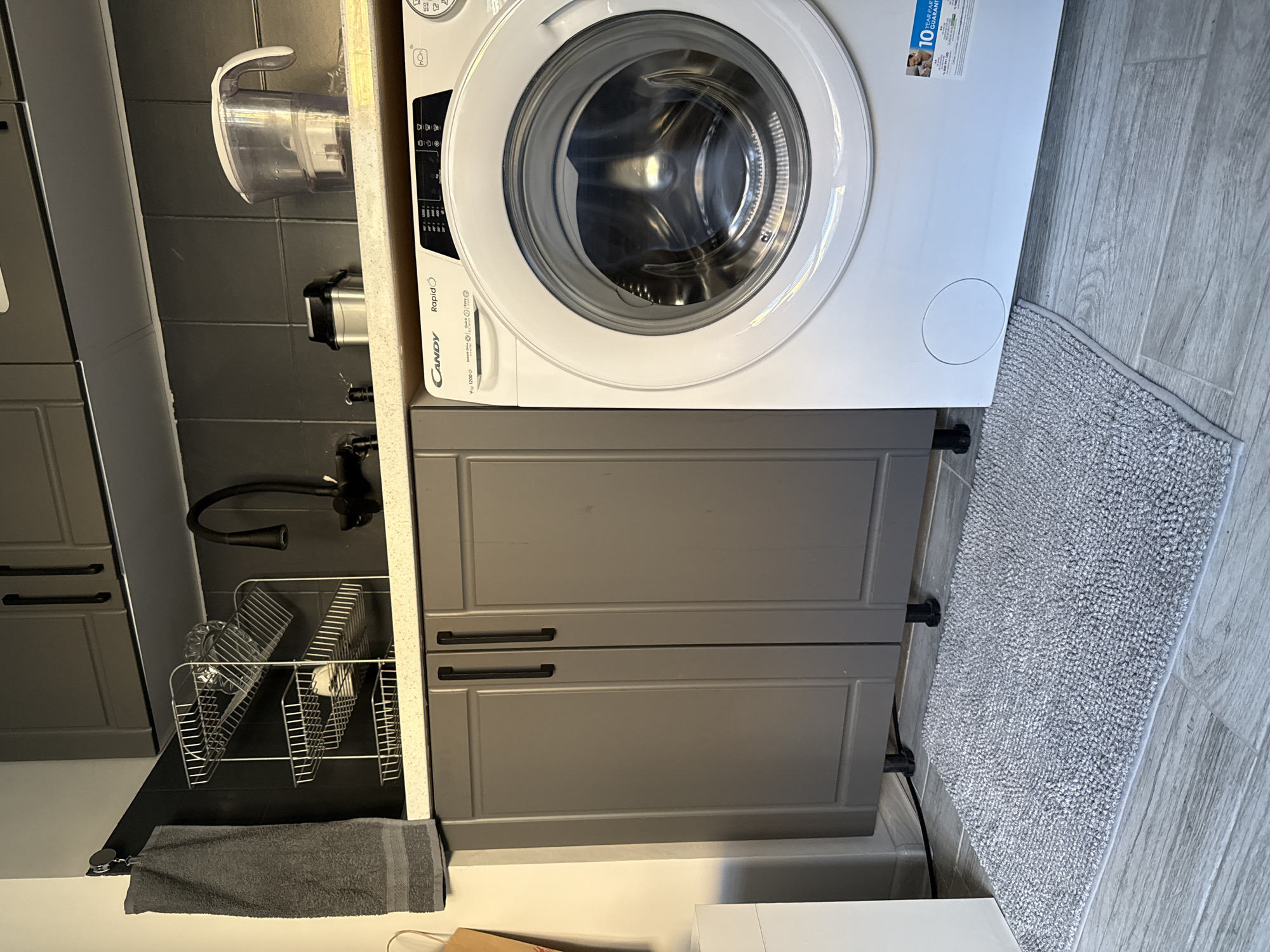 The image depicts a compact laundry area, likely in an apartment, situated in Stara Zagora, Bulgaria. The primary focus is a white Candy RapidO washing machine positioned on a tiled floor. To the left of the washing machine, a set of dark gray cabinets with sleek handles are visible. Above the cabinets, a gray tiled backsplash extends to the ceiling. A chrome dish rack with several items, including glassware, sits on the countertop. A dark gray towel hangs from a hook on the wall to the left, next to the cabinets. A small, textured gray rug is placed in front of the washing machine and cabinets. The background wall is dark gray tile. A water filter pitcher is visible on the shelf above the washing machine, next to a small metallic trash can. The lighting suggests an indoor setting with artificial light, possibly from overhead fixtures.