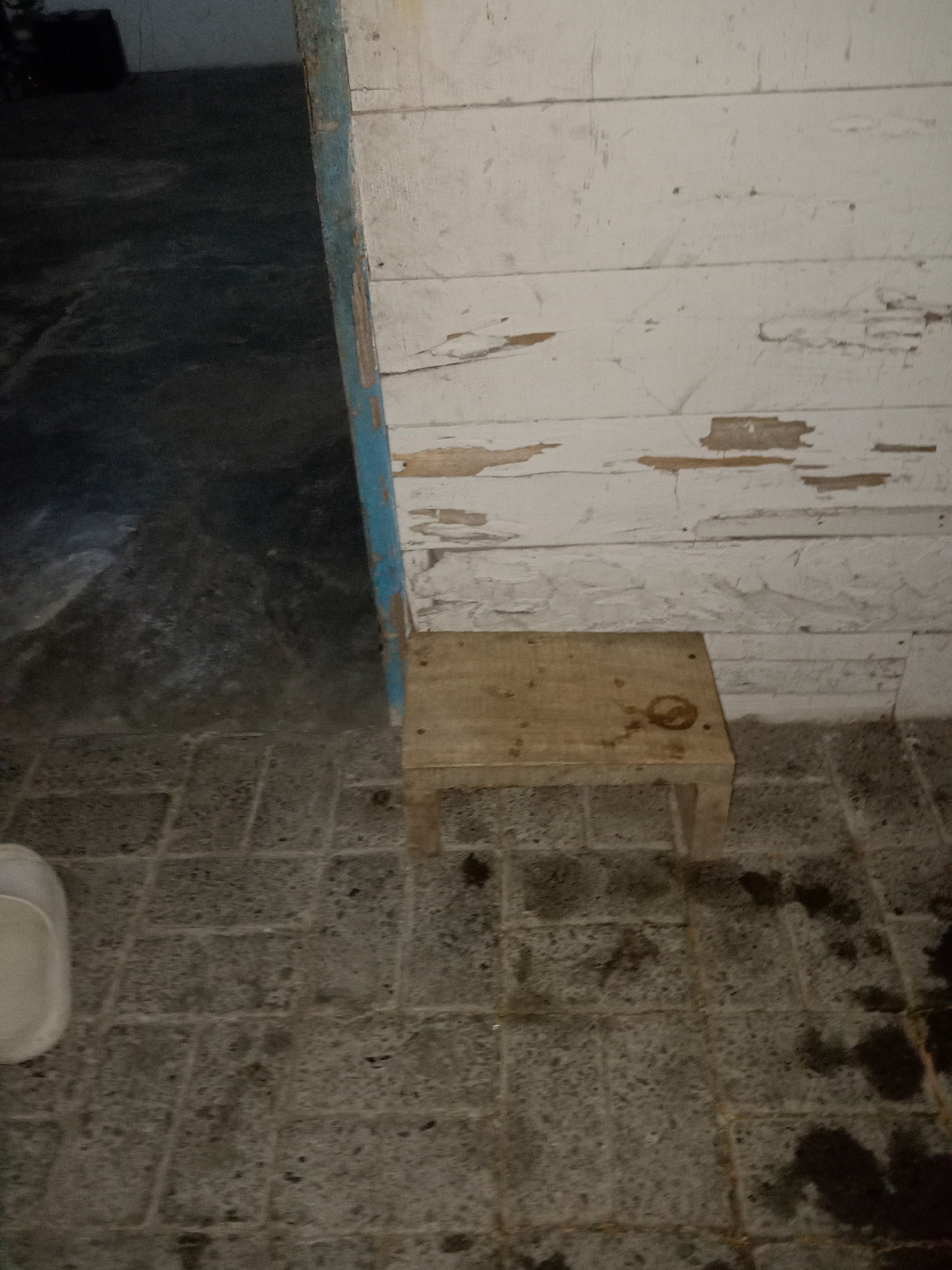 The image depicts a wooden stool placed on a tiled floor in front of a weathered, white wooden wall. A blue and brown painted door frame is visible to the left of the stool. The environment appears to be outdoors, possibly a patio or alleyway. There are dark stains on the floor around the stool, and some of the paint is peeling from the wall. The overall scene is dimly lit, suggesting it might be evening or an overcast day. The location is identified as Jumput, Indonesia.