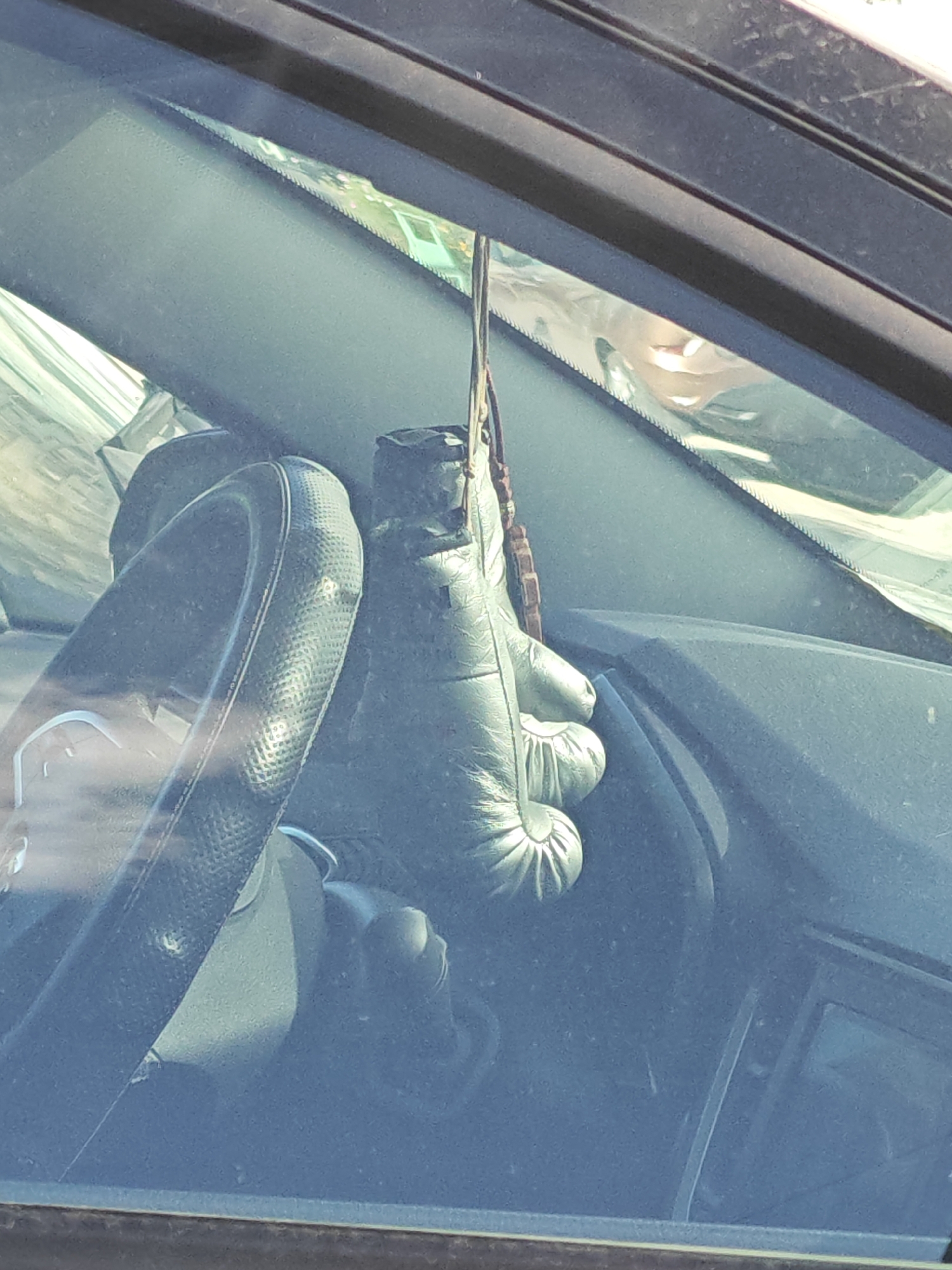 The image shows the interior of a car, viewed through the windshield. A boxing glove is hanging from a string, likely as an air freshener or decoration, from the rearview mirror area. A portion of the steering wheel is visible on the left side of the frame. The dashboard and parts of the car's interior are also visible. The scene appears to be outdoors, with sunlight reflecting off surfaces and visible in the windshield. The context of Tolyatti, Russia is not visually ascertainable from the image itself.