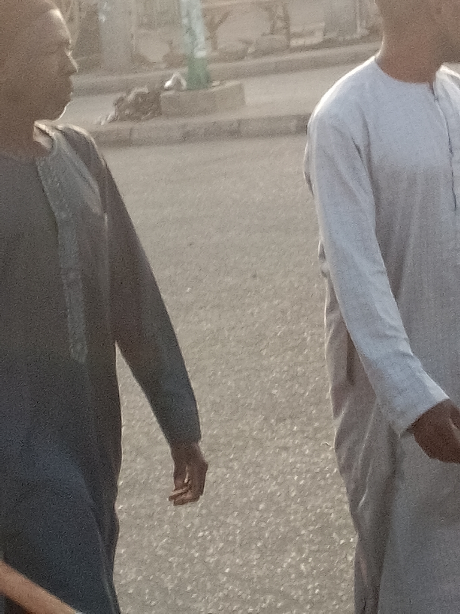 Two individuals, possibly male, are walking on a paved road in Pantame, Nigeria. The person on the left is wearing a dark, long-sleeved garment with a patterned neckline, and their left arm is visible. The person on the right is wearing a light-colored, long-sleeved garment. The background shows a raised concrete curb and a dusty, outdoor environment. The lighting suggests it is daytime, possibly late afternoon, given the shadows and overall light quality. The image is out of focus, hindering precise identification of facial features or finer details of the clothing and surroundings.