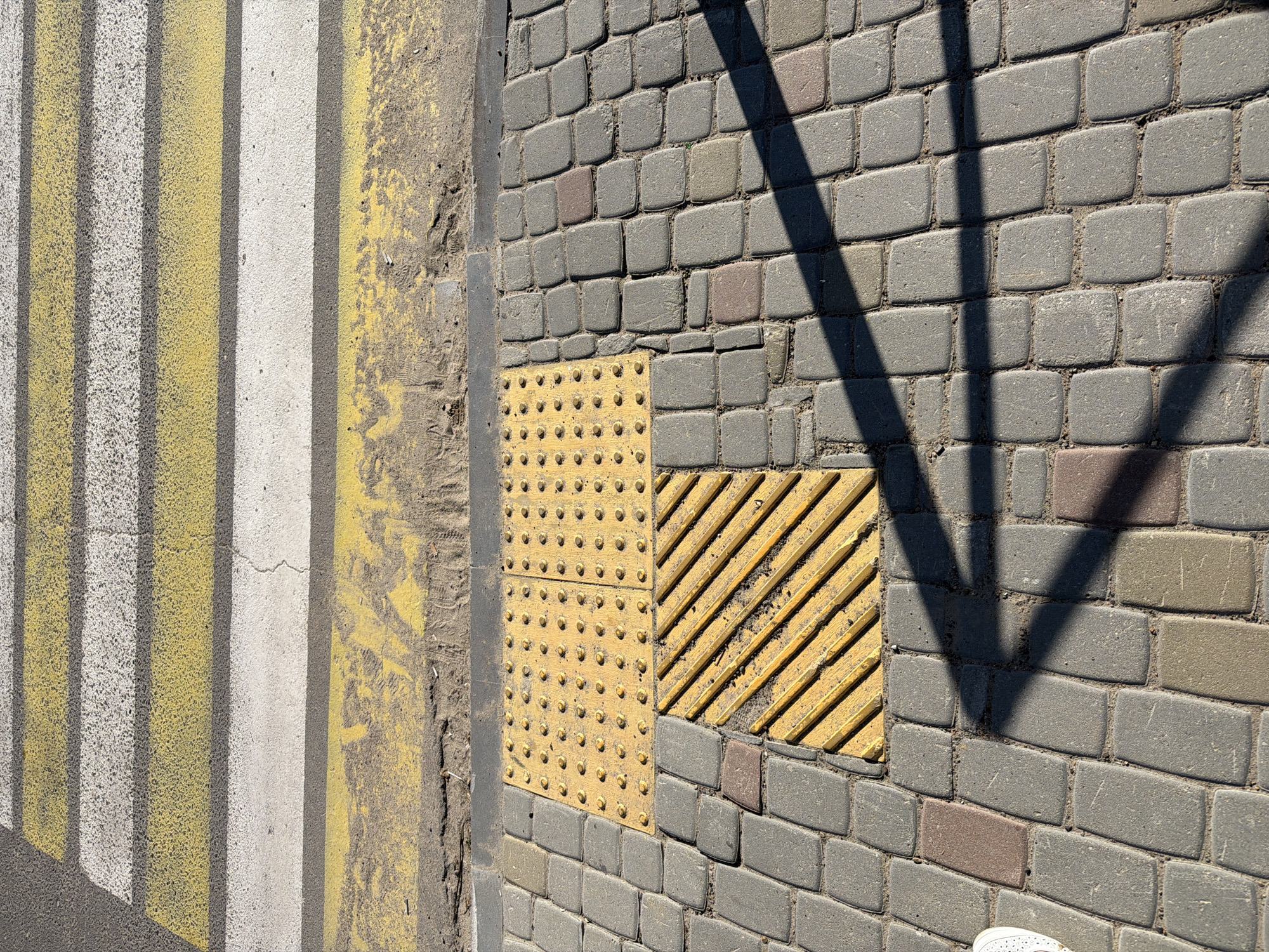The image shows a close-up view of a pedestrian crossing and tactile paving on a city sidewalk in Okha, Russia. The pedestrian crossing features alternating white and yellow stripes painted on asphalt. Adjacent to the crossing, the sidewalk is paved with grey interlocking stones. Part of the sidewalk has yellow tactile paving, including a section with raised domes and another section with raised diagonal lines. The scene is illuminated by sunlight, casting a distinct shadow across the cobblestones. A small portion of a white shoe is visible on the far left of the image, suggesting a person is present but not fully in frame.