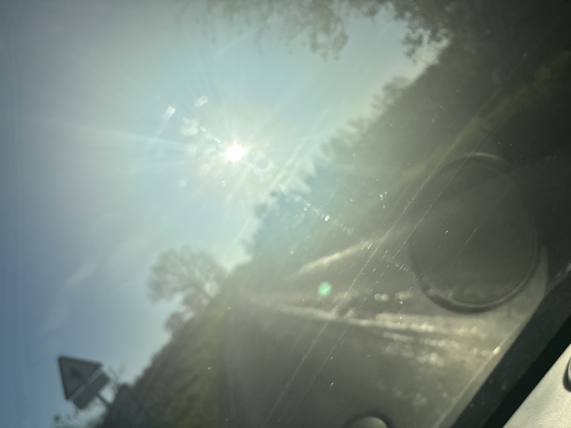 The image appears to be taken from inside a vehicle, looking through the windshield. The sun is shining brightly, causing significant glare and obscuring the view. A road is visible in the distance, flanked by trees. A road sign, triangular in shape, can be seen on the left side of the frame. The presence of a road sign suggests that the vehicle is located near or within Ruyton-XI-Towns, United Kingdom. The exact location cannot be determined without further details.