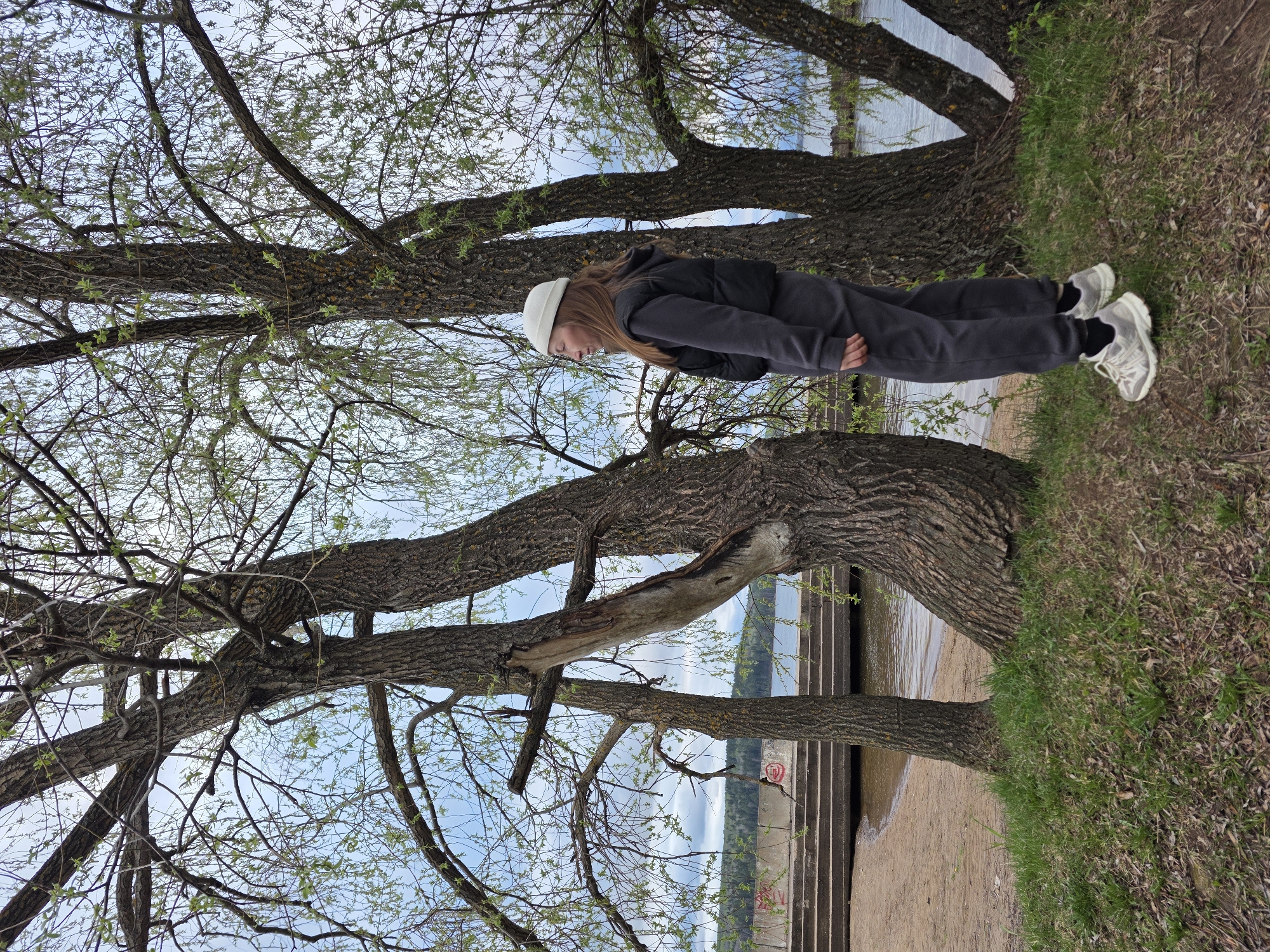 A full-body shot depicts a young woman outdoors near a body of water, likely the Kama River, in Chaikovsky, Russia. She wears a white beanie, dark gray two-piece suit, a black vest, and white sneakers. She stands on the grassy edge of a bank, leaning against the side of a large tree. The tree's branches spread across the upper left portion of the frame, partially obscuring a view of the sky and water. In the background are the concrete steps near the shoreline.