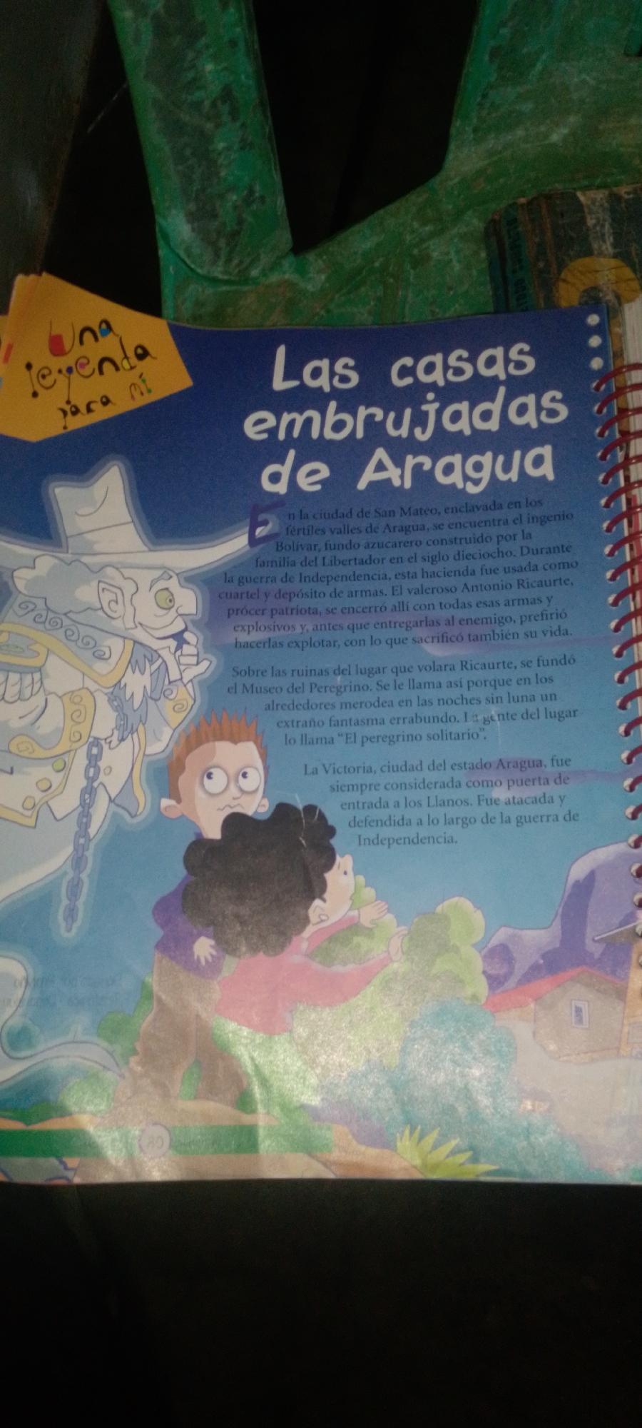 The image is a close-up, slightly angled shot of an open book with a colorful illustration and text. The illustration features cartoonish figures, including a tall, ethereal white figure with a hat and chains, and two children looking surprised or scared. The children are in the foreground, with one reaching out towards the white figure and the other holding onto the first. Behind them are trees and a stylized building. The text on the page is in Spanish and appears to be a story or historical account, with the title "Las casas embrujadas de Aragua" (The haunted houses of Aragua) prominently displayed. A yellow sticky note with the handwritten text "Una leyenda para mí" (A legend for me) is attached to the upper left corner of the book cover. The overall tone of the illustration is whimsical and a bit spooky, suggesting a children's book or educational material. The provided context of La Tagua, Venezuela, is not directly visible in the image itself but could be related to the content of the book.