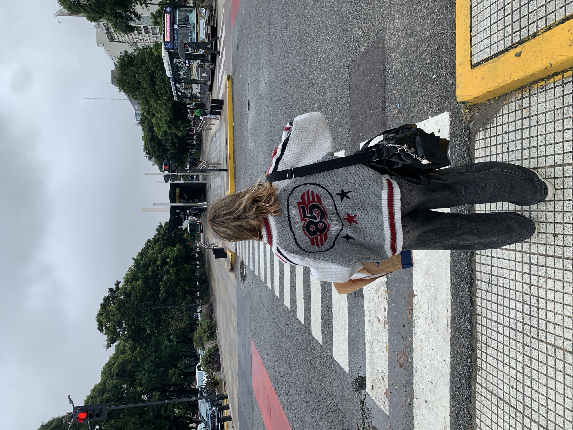 A young person with long, wavy blond hair is shown from the back as they cross a street on a zebra stripe crosswalk. The individual is wearing a gray sweater with red and white stripes, and a large "58" graphic with stars. They are also wearing dark gray or black pants and carrying a black bag with a shoulder strap.
The environment appears to be an urban setting in Buenos Aires, Argentina. Buses and cars are visible in the background, along with trees lining the street and buildings. The weather seems overcast, with an even, gray sky. The crosswalk is marked with alternating white and gray stripes, and red paint on the road surface. The curb on the right has yellow paint.