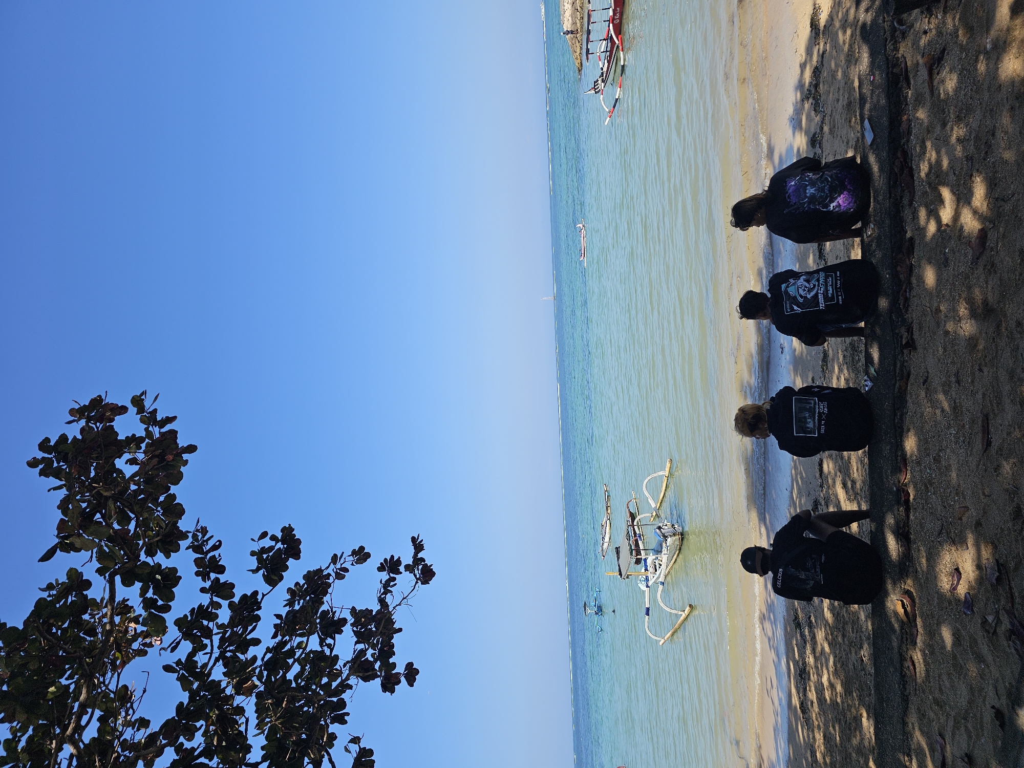 Four people sit on the sand, facing the ocean under a tree. The people are dressed in dark t-shirts. The ocean is calm with several boats on it, including a traditional Balinese fishing boat in the foreground. The sky is clear and blue. The scene is likely a beach in Denpasar, Indonesia, known for its beautiful coastline and traditional boats.