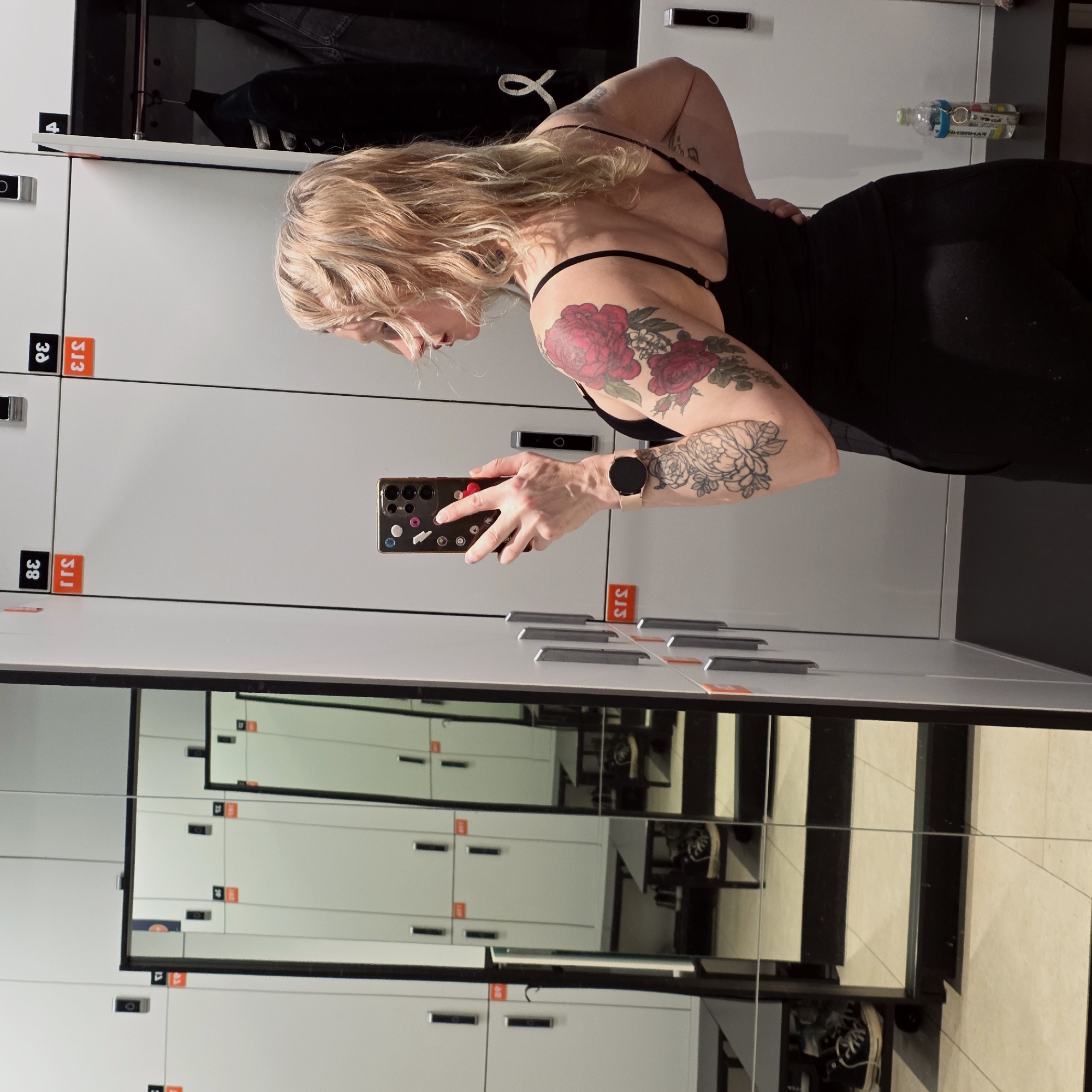 The image is taken indoors, presumably in a locker room or changing area, as indicated by the rows of lockers. A person is visible in the frame, holding a smartphone and seemingly taking a selfie or a photograph of themselves. The person is wearing a dark-colored, form-fitting garment, possibly activewear, and has fair skin, blonde hair, and tattoos on their arms. The person is wearing a smart watch.
The background consists of light-colored lockers with visible numbering and handles. A partial reflection in the mirrors shows additional rows of lockers and flooring with a black and white design. The environment looks clean and organized. There's a water bottle on top of the lockers and clothes hanging inside.
The image does not provide sufficient information to determine whether the scene is located in Saint Petersburg, Russia.