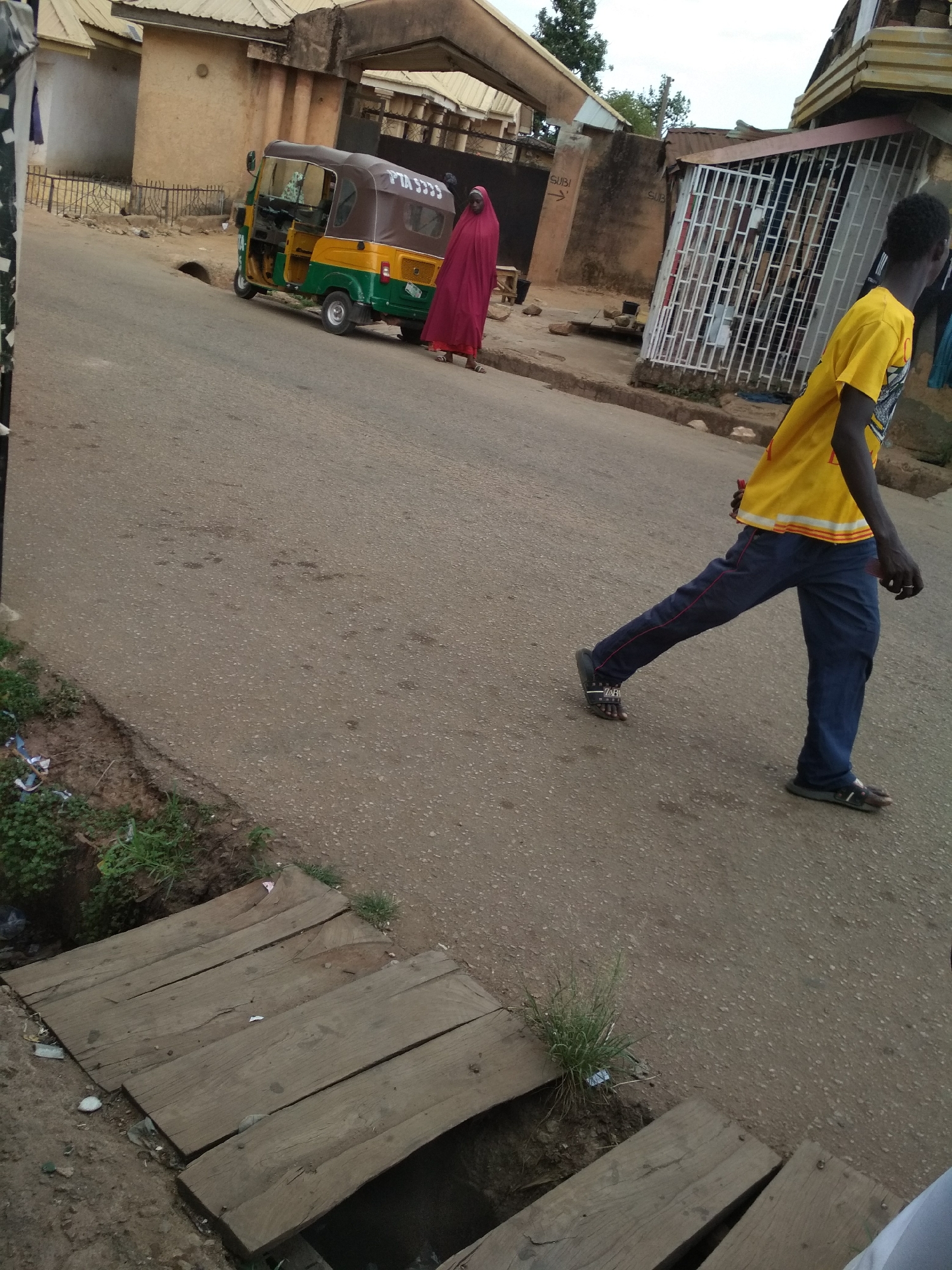 The image shows a street scene in Jos, Nigeria. An adult person wearing a yellow shirt and blue pants walks on the right side of the frame. A "keke napep" (tricycle taxi) with a green and yellow body and brown roof is parked near a person in a red robe. A covered drainage ditch is visible in the foreground. Buildings are present on both sides of the street.