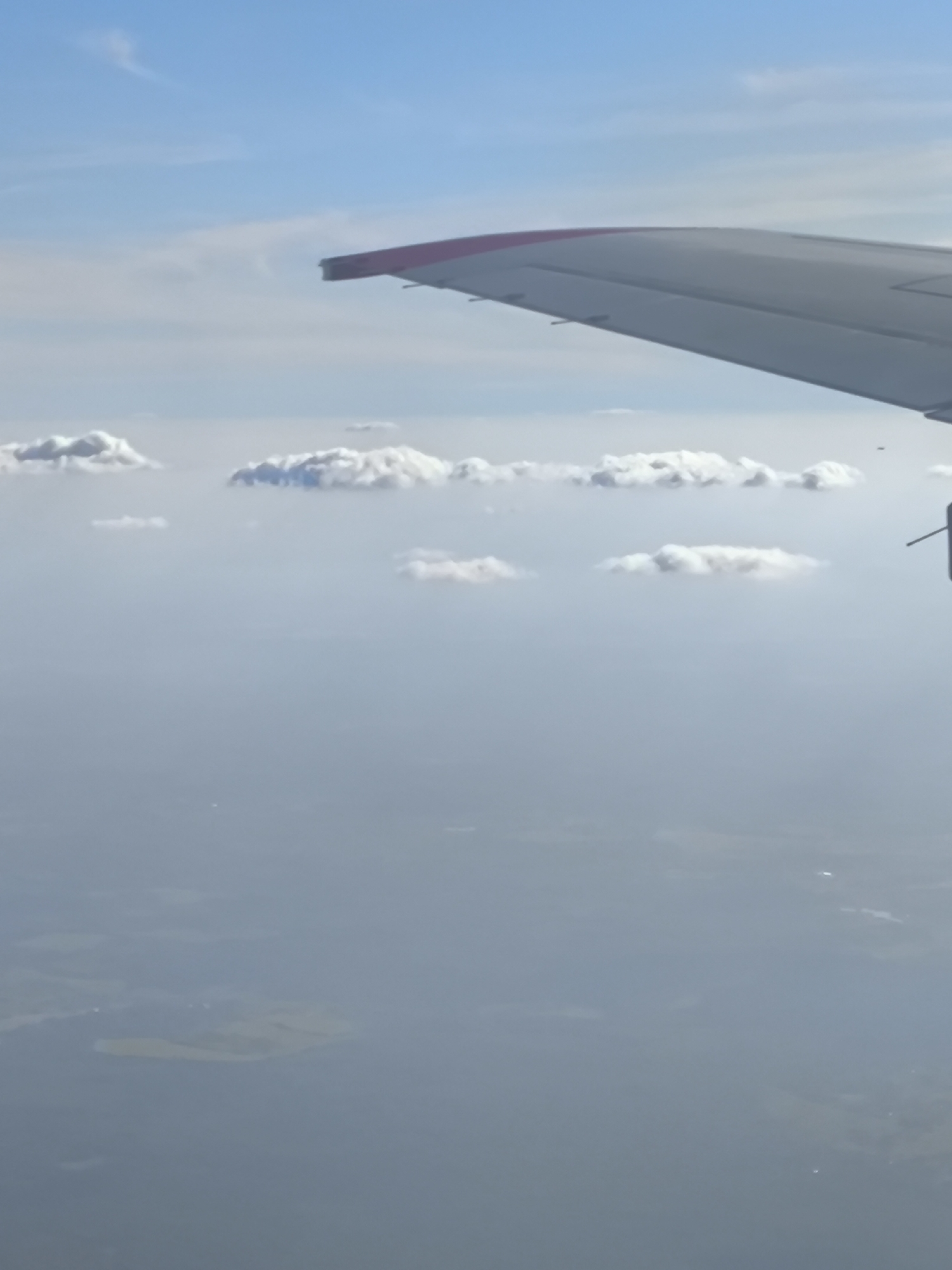 The image depicts an aerial view from an aircraft, with the right wing visible in the upper-right quadrant. The sky is clear blue at the top, transitioning to a lighter, hazy blue further down. Scattered white cumulus clouds are present in the mid-ground, floating above a dense atmospheric haze. Below the haze, a vast expanse of water and land is visible, heavily obscured by distance and atmospheric conditions. A distinct, irregularly shaped yellowish landmass, likely an island or part of a coastline, is discernible in the lower-left. No individuals are visible. The specific geographical location in the city of Unknown, Unknown, cannot be determined from the visual information provided.