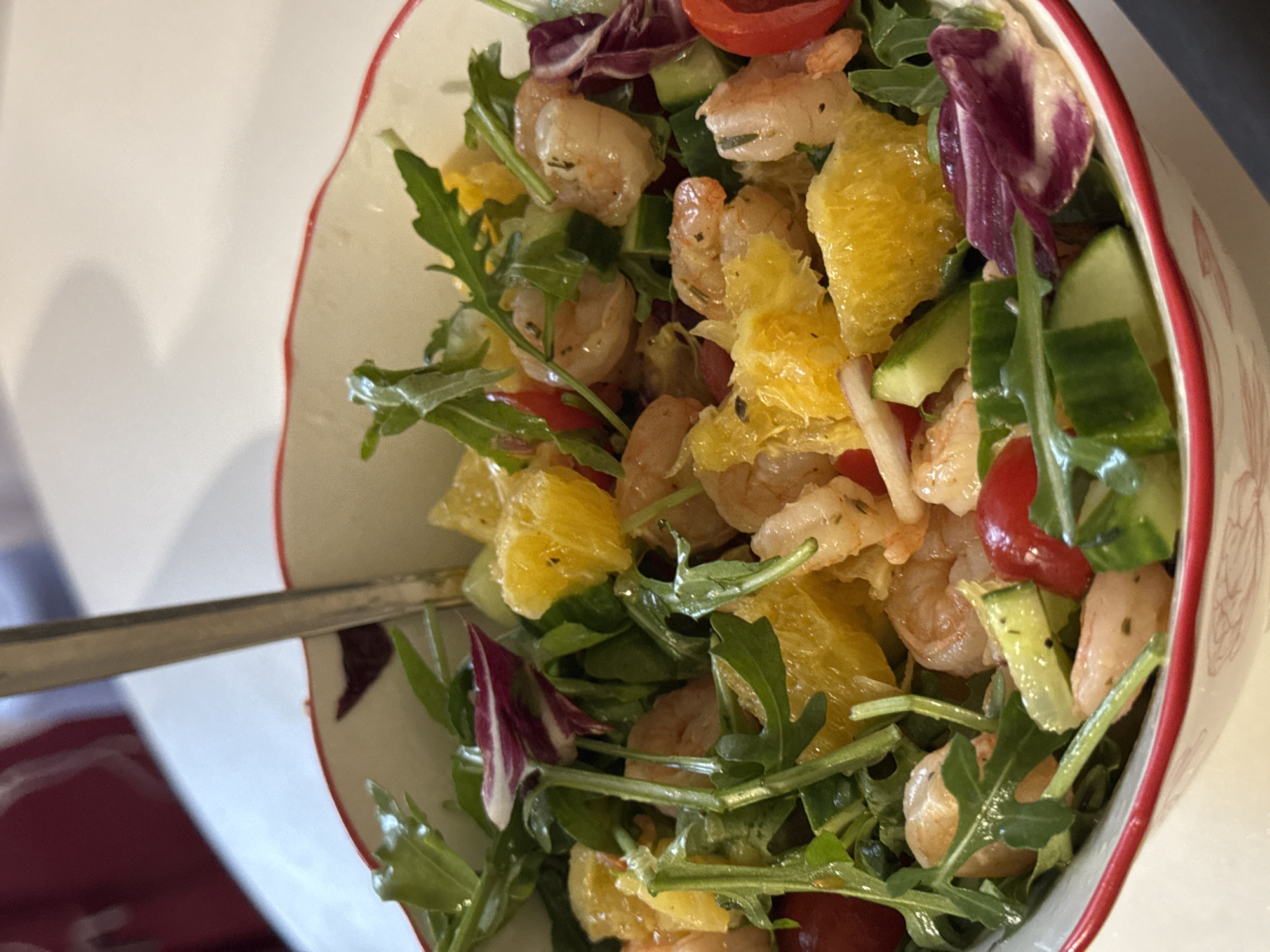 A close-up shot depicts a colorful salad in a bowl. The salad includes shrimp, segments of orange, arugula, cucumber, and halved cherry tomatoes. A silver fork is partially visible in the bowl. The bowl itself is white with a red rim. The scene suggests an indoor setting with a bright, possibly natural, light source. There is no information about the location in the city of Chaikovsky, Russia.
