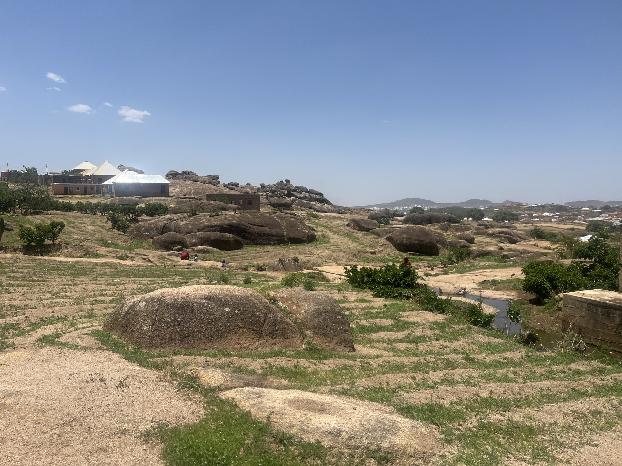 The image shows a landscape in Jos, Nigeria, characterized by large rocky formations and sparse vegetation under a clear blue sky. In the background, there are buildings with tin roofs and surrounding trees, indicating human settlements. Several people are scattered across the area near the center of the frame. A small body of water (possibly a stream or ditch) is visible on the right side of the frame, close to a concrete structure. The foreground is dominated by light brown soil interspersed with patches of grass.