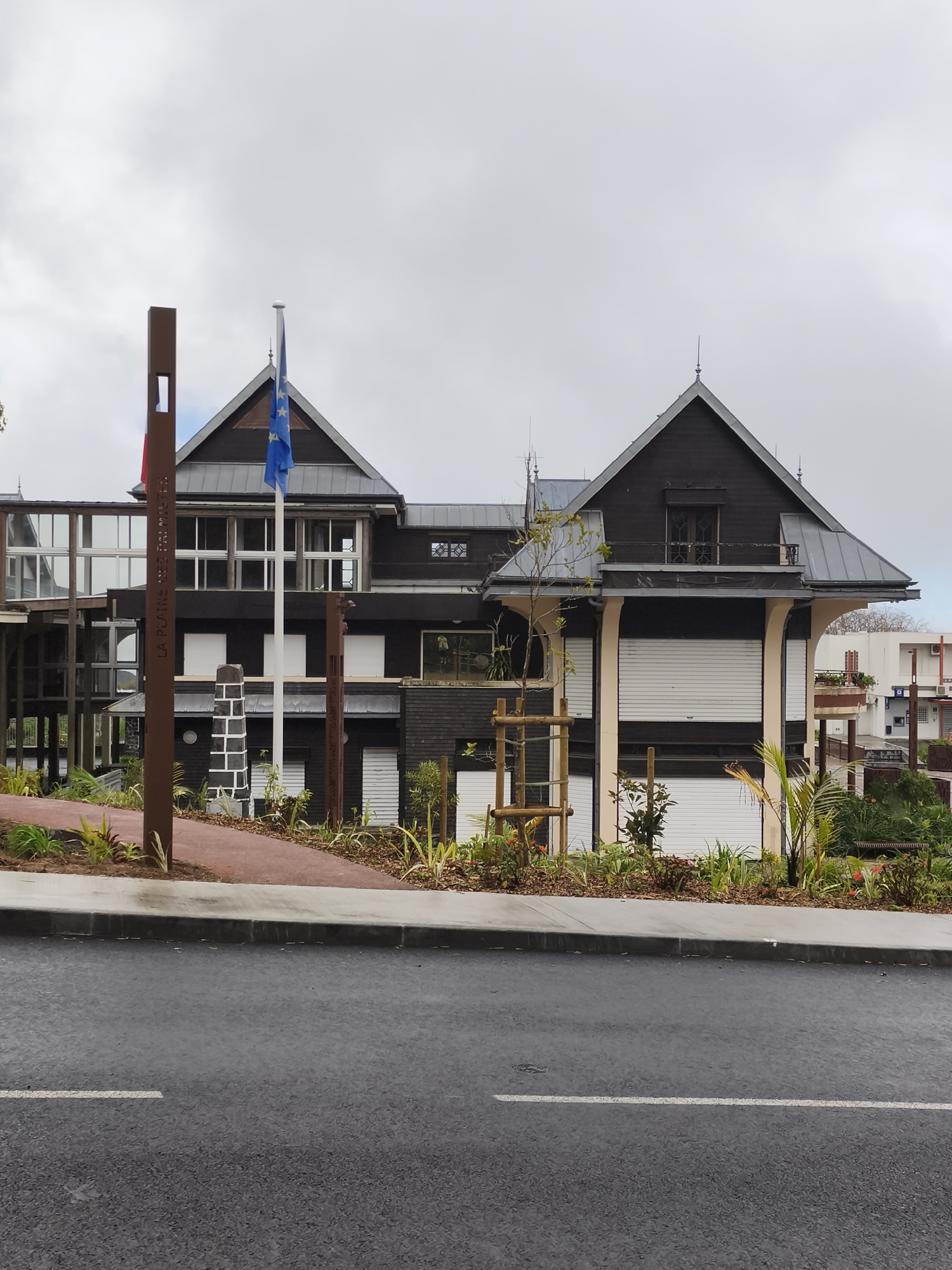 The image depicts the entrance to a municipal building in La Plaine-des-Palmistes, France. The building, constructed with dark wood and light-colored trim, features multiple gabled roofs and a modern, multi-story glass-paneled walkway connecting different sections. Flags, including the European Union flag, are visible near the entrance. A tall, brown metal sign with text is positioned to the left of the building, and a red pathway leads towards it. The area is landscaped with various plants and greenery. The scene is captured from a street-level perspective, with a section of asphalt road and sidewalk in the foreground. The sky is overcast with clouds. There are no people visible in the image.