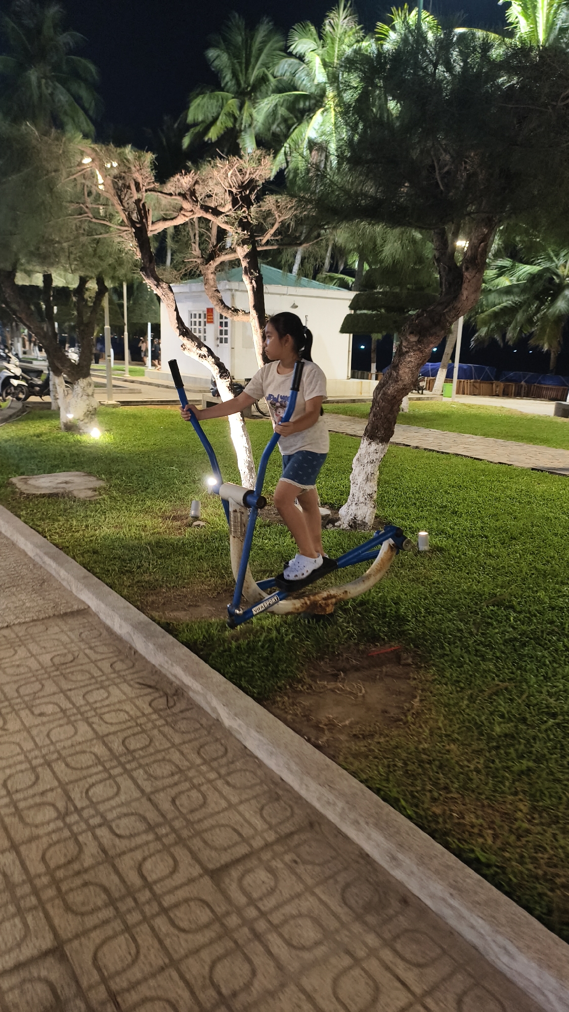 In Nha Trang Ward, Vietnam, at night, a young girl with dark hair tied in a ponytail is using an outdoor elliptical exercise machine. She is wearing a light-colored t-shirt and blue denim shorts. The elliptical machine is blue and white. The girl is standing on a paved walkway with a patterned design, and behind her is a well-maintained grassy area illuminated by outdoor lighting. Several trees with thin trunks and sparse foliage are scattered around the space. In the background, a white building with windows is visible, along with more palm trees. The overall setting appears to be a park or recreational area.