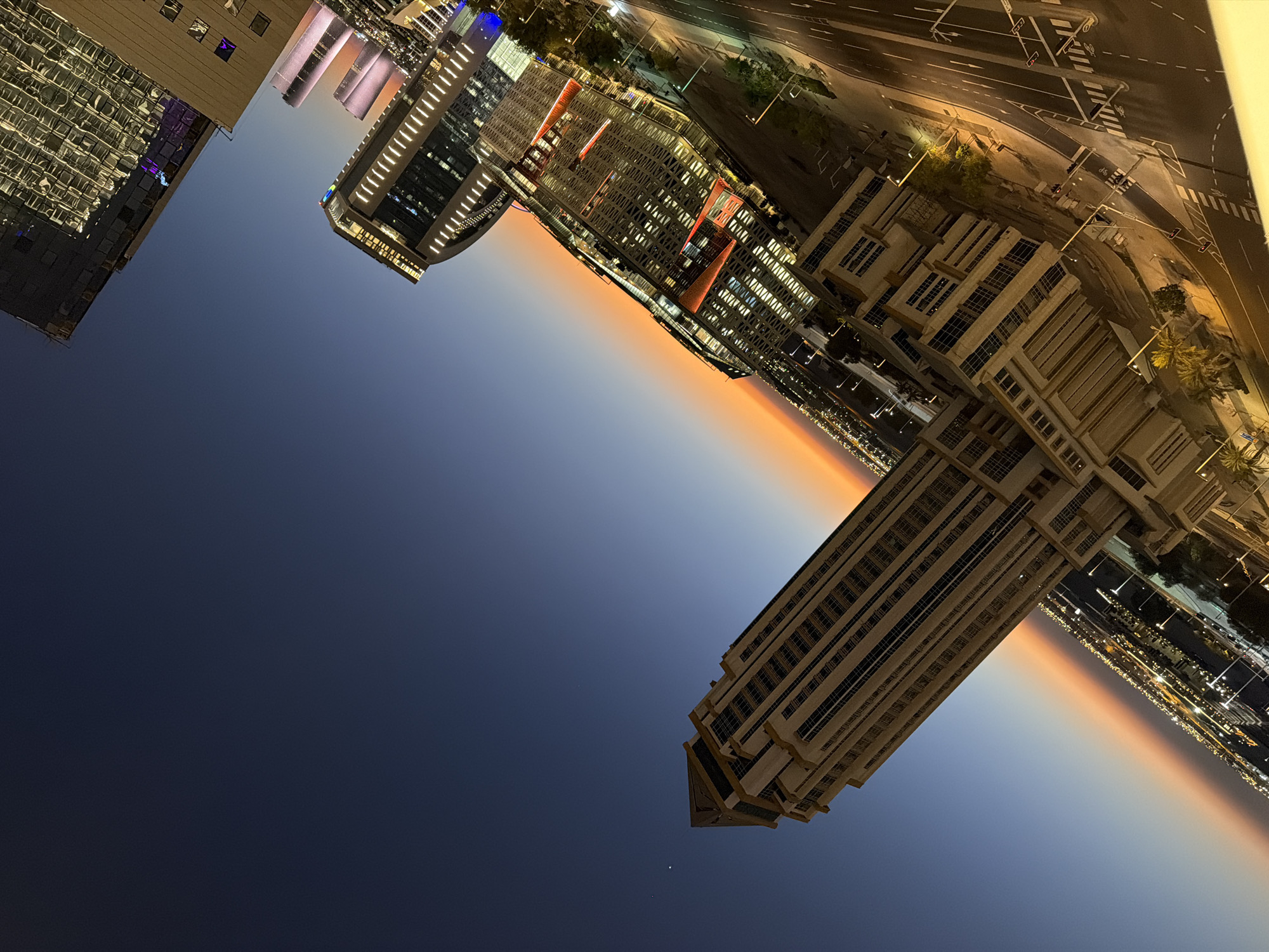 An angled night time shot showcases a city skyline at dusk with a gradient of orange and dark blue in the sky. Dominating the foreground is a tall, beige, tiered skyscraper with many windows, tilted on its side. To the right, other modern buildings are visible, some with illuminated windows and colorful exterior lighting. Roads and streetlights are also present, indicating an urban environment. A faint star is visible in the dark blue portion of the sky.