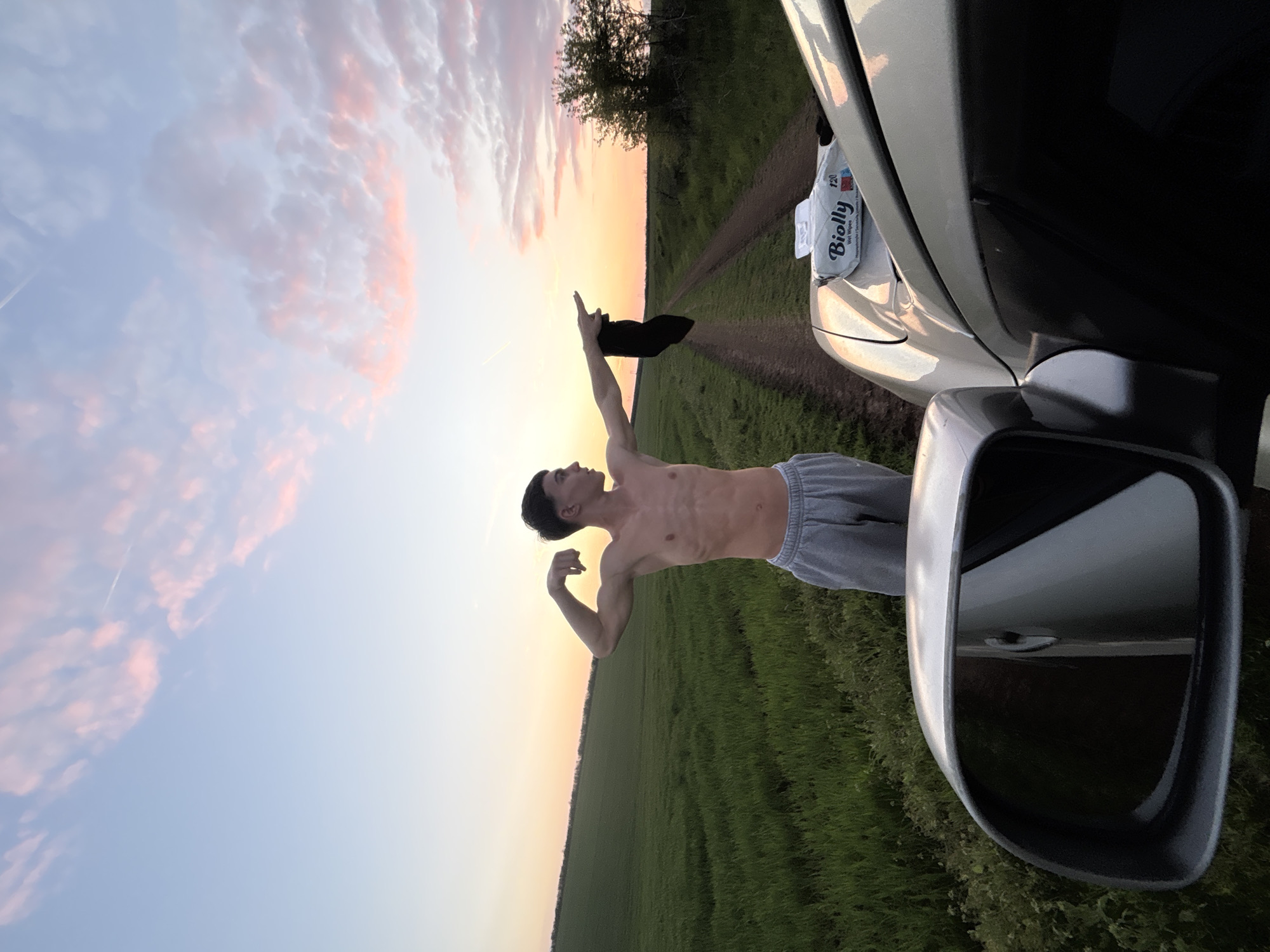 A shirtless male stands beside a silver car, on a dirt road amidst a green field at sunset. He holds a black hat aloft with his right hand and flexes his left arm. A package of "Biolly" wet wipes sits on the car's hood. The photo appears to be taken near Lehchevo, Bulgaria.