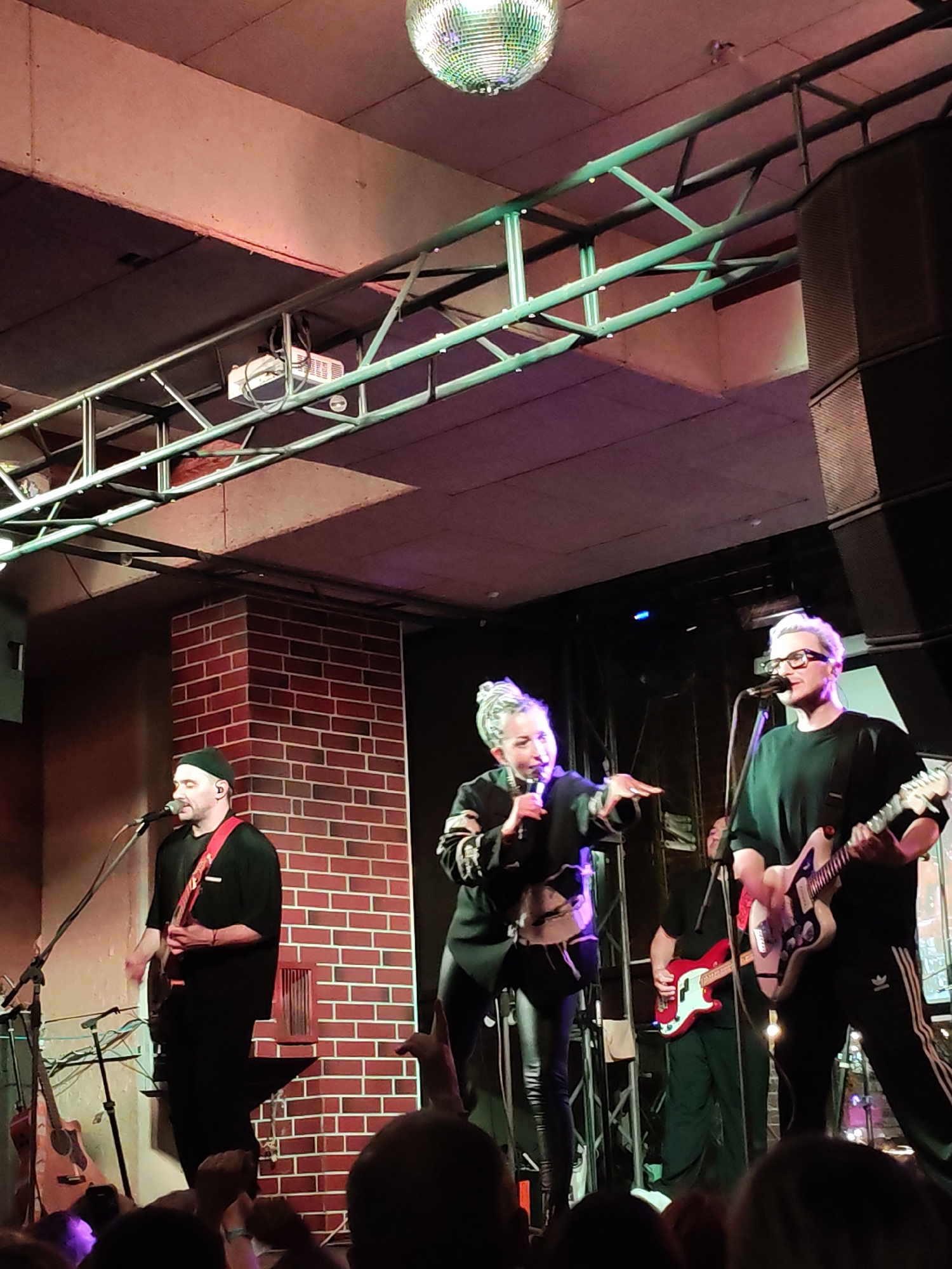 This image captures a live music performance by a band on an indoor stage, likely in a club or concert venue in Izhevsk, Russia. The setting features a prominent metal truss structure overhead, illuminated with green light, and a large disco ball hanging from the ceiling. A brick wall forms part of the stage background on the left.
Three main performers are visible on stage. In the center, a female vocalist with light-colored hair, styled upwards, is actively singing into a microphone and gesturing with her right hand. She wears a dark jacket or top over dark pants. To her right, a male guitarist and vocalist with short, light-colored hair and glasses is singing into a microphone while playing a light-colored electric guitar. He wears a dark long-sleeved shirt and dark pants, which feature the "Adidas" three-stripe logo on the side. On the far left, another male guitarist and vocalist, wearing a dark beanie, dark shirt, and a colorful guitar strap, is playing an electric guitar and singing. Behind the central vocalist, a fourth band member is partially visible, playing a red bass guitar. An acoustic guitar rests on a stand on the left side of the stage.
Various stage equipment is present, including microphones on stands, speaker cabinets, and a projector suspended from the truss. The performers appear engaged and energetic, suggesting an exciting live show. The bottom portion of the image reveals the top of several audience members' heads and raised hands, indicating their presence and enjoyment of the event. One audience member is holding a phone, possibly recording the performance. The overall lighting suggests an evening or nighttime event.