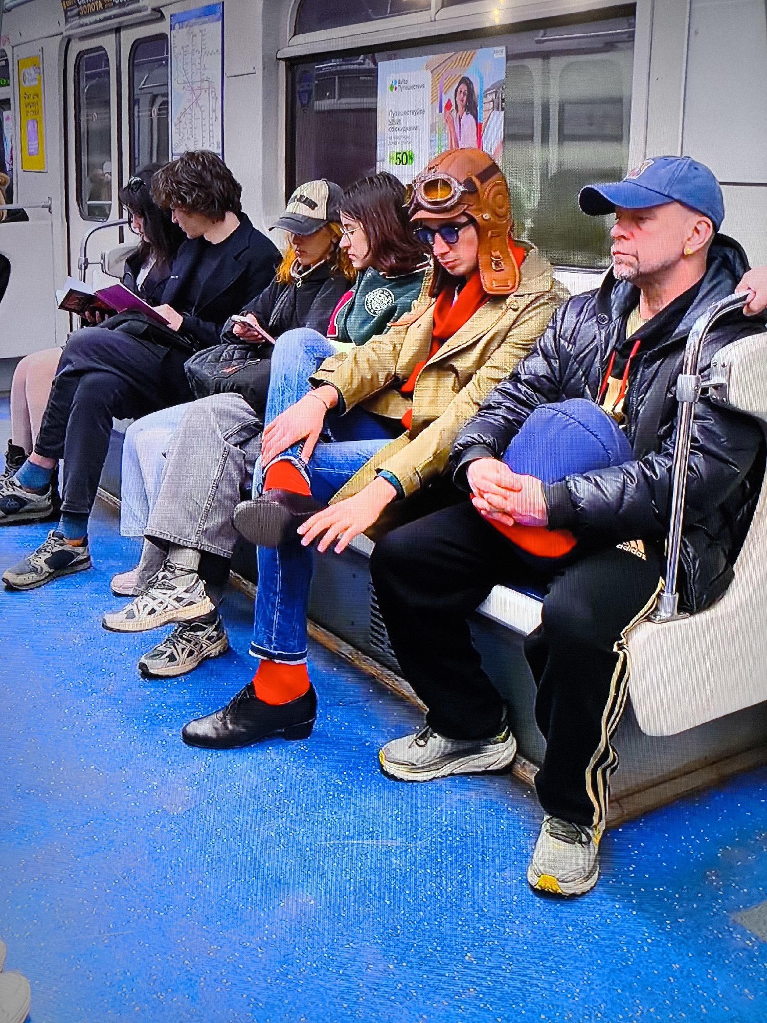 A photograph captures a diverse group of people riding the subway in Odesa, Ukraine. Seated on a subway train, passengers exhibit varied attire and activities. A woman reads a book. A man wears an aviator helmet and sunglasses. Another man wears a blue baseball cap. The subway car features posters on the wall and a blue floor with speckled patterns.