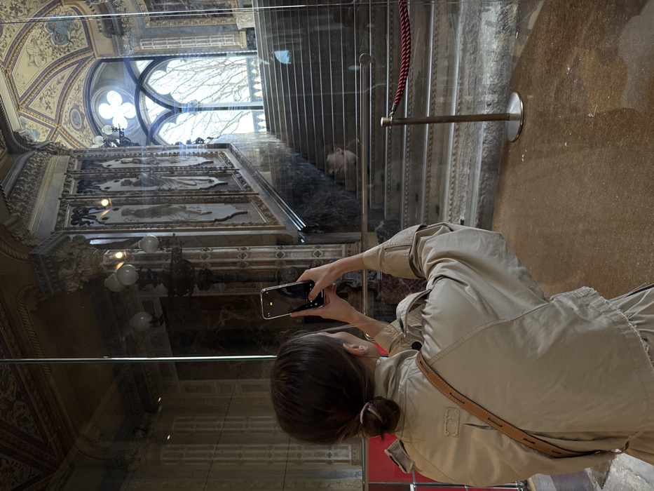 An individual with dark brown hair, tied in a low bun, is depicted from behind, centered in the foreground, wearing a beige or light tan long-sleeved jacket. A brown leather bag strap crosses their right shoulder. They hold a silver-colored smartphone horizontally with both hands, positioned at eye level, appearing to photograph or record the grand interior space beyond a transparent glass barrier.
The scene beyond the barrier reveals a magnificent, ornate marble staircase ascending towards a large, multi-lobed Gothic-style window. The walls flanking the staircase are adorned with intricate decorative paneling featuring figures or patterns, columns with elaborate capitals, and ornate multi-bulb light fixtures. A large, light-colored urn or vase rests on a landing of the staircase. Through the Gothic window, the bare branches of trees against an overcast sky are visible. A red braided rope barrier, supported by a brass stanchion, extends into the frame from the right side, immediately behind the glass barrier. The floor in the foreground is a polished, mottled brown surface. The glass barrier creates reflections, superimposing elements from the immediate foreground onto the background architectural details.
This interior is identifiable as the grand staircase of the **Ca' d'Oro (Palazzo Santa Sofia) in Venice, Italy**, specifically within the Giorgio Franchetti Gallery.