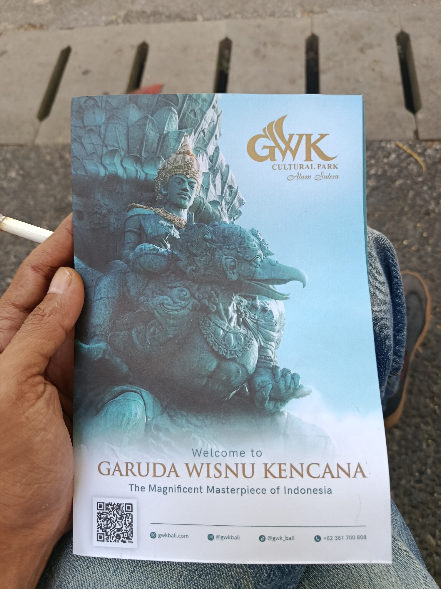 The image shows a close-up of a brochure for Garuda Wisnu Kencana Cultural Park, located in Kuta Selatan, Indonesia. The brochure features a large image of a statue of Garuda and Vishnu. A person's hand is holding the brochure, with a lit cigarette visible in the fingers. The background shows a gray, textured surface, likely pavement. The lower part of the brochure displays the park's name, "GARUDA WISNU KENCANA," and the tagline "The Magnificent Masterpiece of Indonesia." It also includes a QR code, website address (gwkbali.com), social media handles (@gwkbali and @gwk_bali), and a phone number (+62 361 700 808). The person is wearing denim jeans.