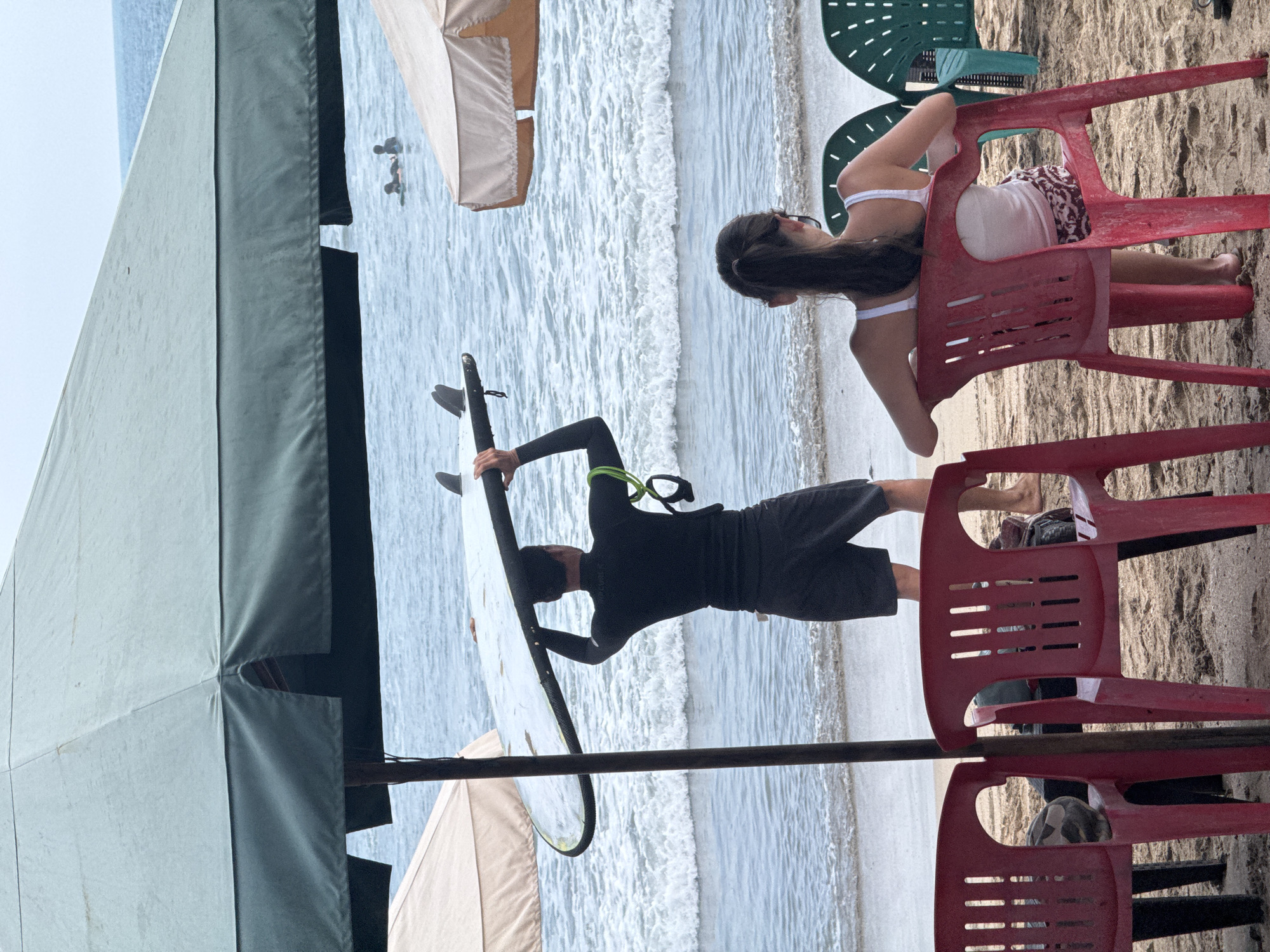 A medium shot captures a beach scene in Kuta, Indonesia, under a large, faded green umbrella. In the foreground, two red plastic chairs are occupied by a woman facing away from the camera, wearing a white tank top and patterned shorts. To her left, a man in a black wetsuit and dark shorts stands holding a surfboard overhead, facing the ocean. The water, with gentle waves, is visible in the background, where several other figures can be seen in the distance, some on surfboards. The beach is sandy, with other chairs and umbrellas also present, suggesting a public beach area. The lighting indicates it is daytime, likely overcast due to the diffused light and the subdued colors.