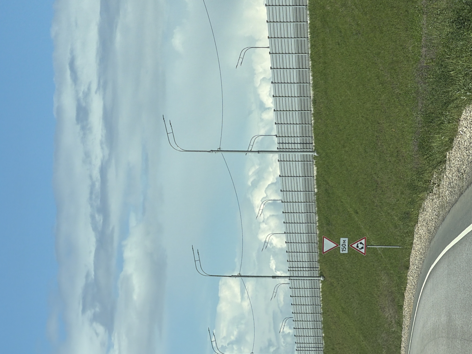 The image shows a scene alongside a road. On the right, a paved road with a white edge marking is visible. Beside the road is a grassy area. A signpost indicates a roundabout 150 meters ahead. Beyond the grass is a barrier, potentially bordering another area. On the left, the sky is visible, featuring a mix of blue sky and white, puffy clouds. Tall metal poles are spaced along the barrier. The city of Zaozerye, Russia is not evident from the image.