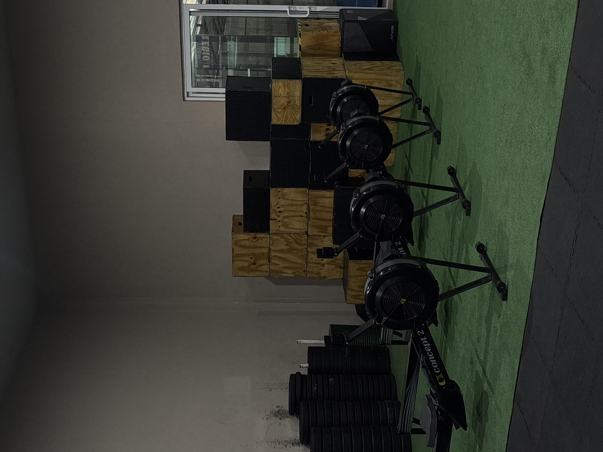 The image depicts an indoor gym or fitness facility, showcasing various workout equipment. In the foreground, three black Concept2 rowing machines are aligned on a green artificial turf floor. The text "Concept 2" is visible on the side of one of the rowers.
To the left of the rowing machines, against a plain light brown or gray wall, there is a stack of plyometric boxes. These boxes alternate between light wooden ones and black ones, some of which feature a textured, dimpled surface. Further to the left, a collection of black weight plates, likely bumper plates, are stacked on the floor.
The floor is predominantly covered by a bright green artificial turf where the rowing machines rest, transitioning to black rubber matting on the right side of the frame. In the upper left corner, a window reflects light and what appears to be reversed text, possibly "LEGIO" or a similar word. Next to this window, on top of some boxes, is a small black box with the white text "6cm.24"" visible on its side. The overall lighting is dim, suggesting an indoor setting with artificial illumination. There are no people visible, and no activity is currently taking place, giving the scene a quiet and prepared atmosphere.