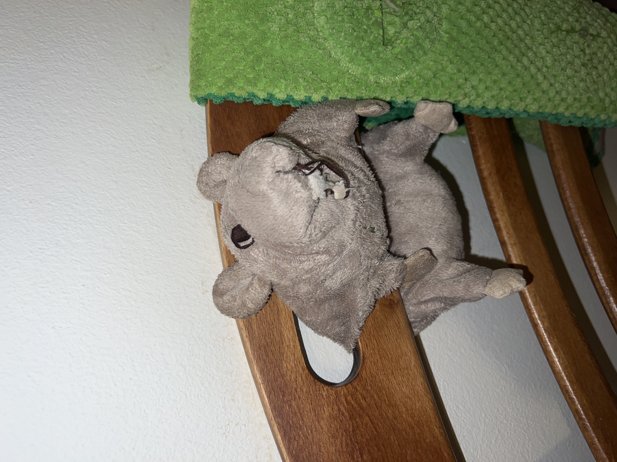 A plush grey animal toy rests on a wooden chair, underneath a green crocheted seat cover. The chair is positioned against a white wall. No people are present in the image. There is no indication of the image being taken in Belgrade, Serbia.