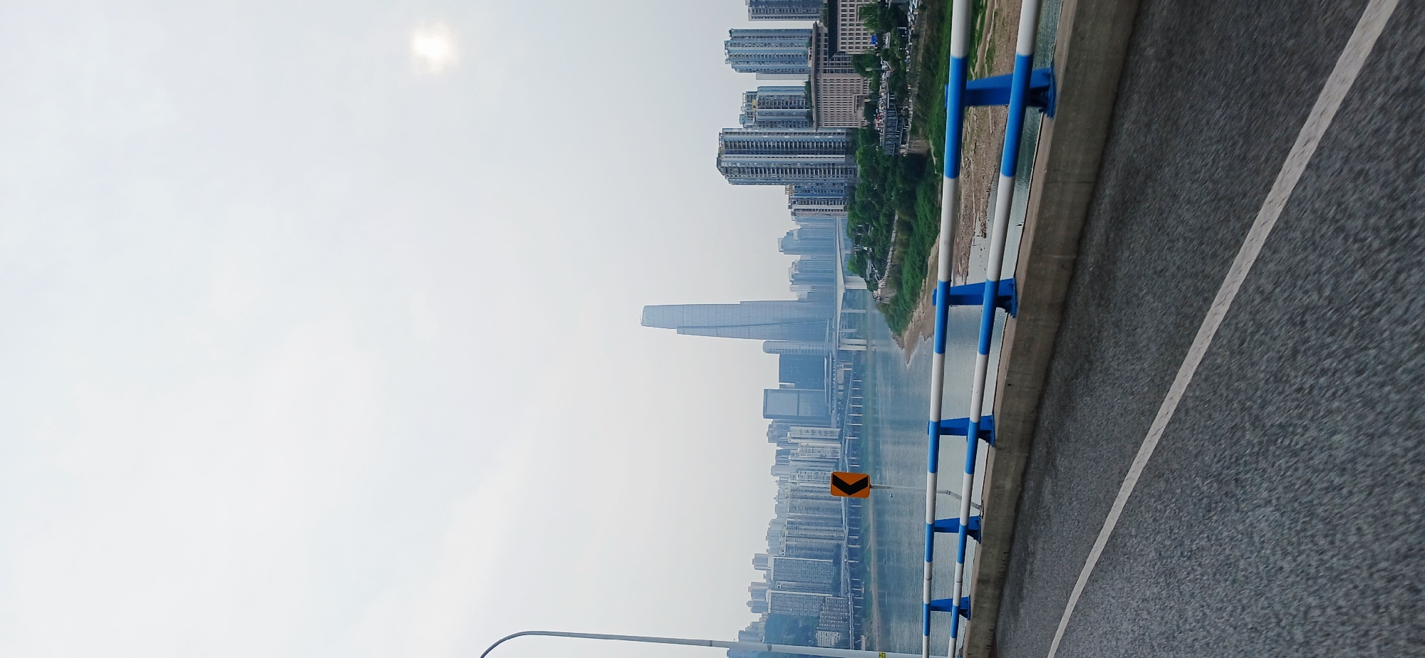 The image shows a view of the city of Yuzhong District, China, from a bridge. A line of high-rise buildings is visible across the water, including a tall, flat-topped skyscraper. The bridge's railing is white with blue supports. The road surface is asphalt with a white lane marker. The sky is overcast.
