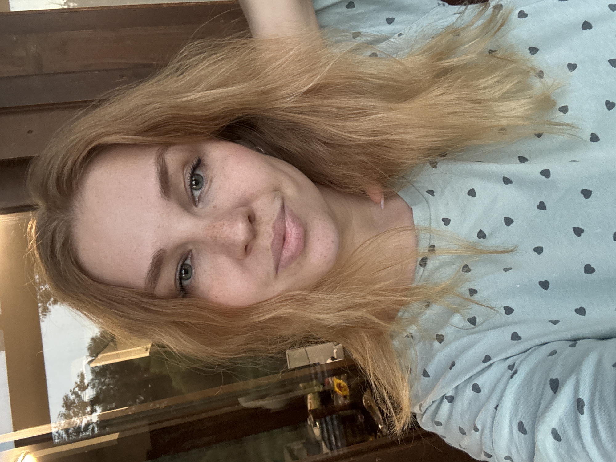 The image is a selfie of a young woman with long, blonde hair and freckles, looking directly at the camera. She is wearing a light blue t-shirt with a pattern of dark gray hearts. Her left hand is raised to her head, and her right arm is resting on her shoulder. The background appears to be indoors, with a window to her left reflecting trees and the sky. To her right, there is a wooden surface. The scene is not identifiable as being in the city of Rusanovo, Russia.