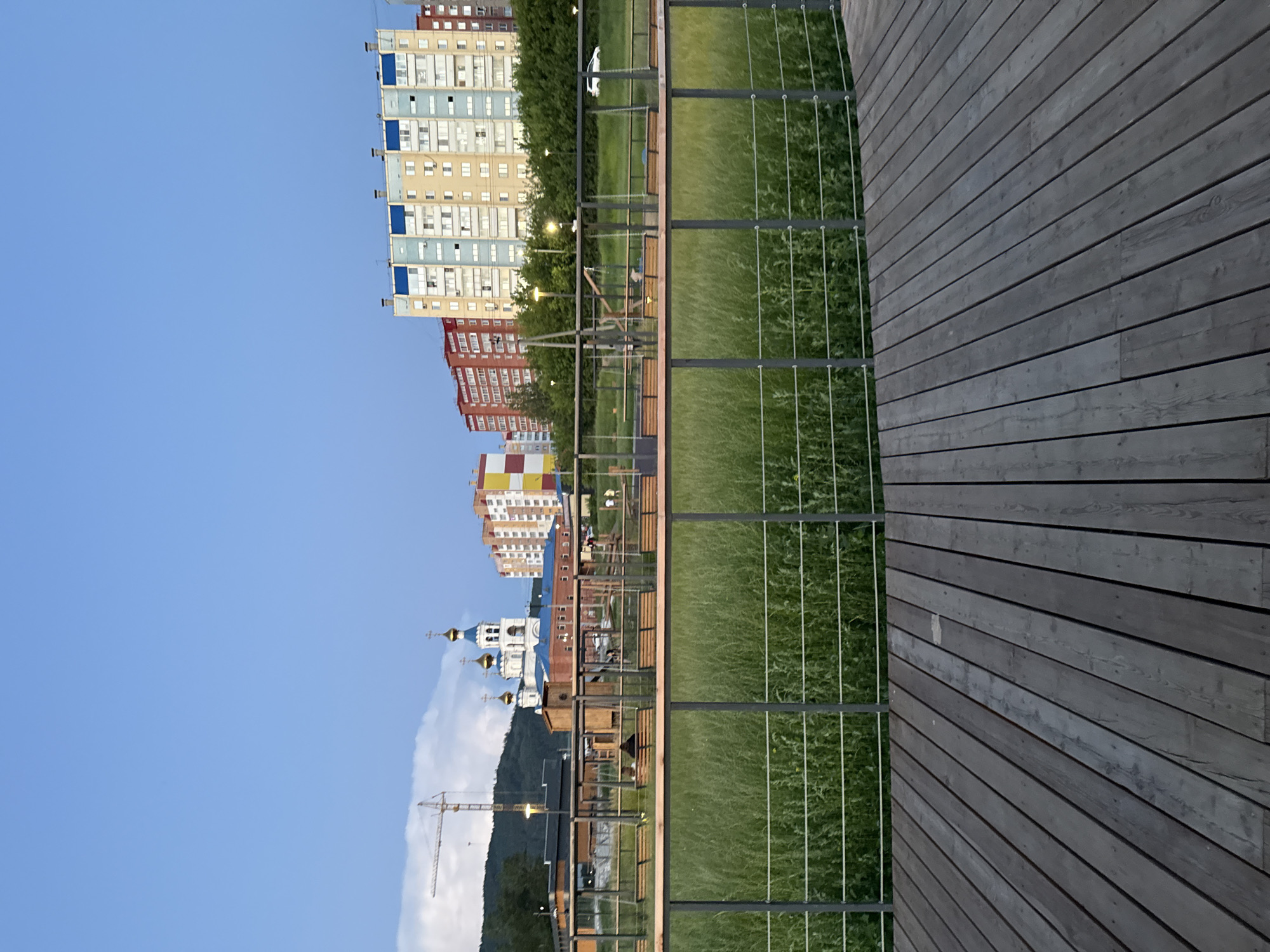 The image captures a scene in Miass, Russia, featuring a wooden deck in the foreground, overlooking a grassy area with benches and swings. Beyond the park, several multi-story buildings are visible, along with a white church with blue domes and a construction crane. The sky is clear and blue, with a hint of sunset colors. Several individuals are present in the park, some seated on benches, others walking or standing near the playground equipment. A white car is parked on the side of the road in the background. The overall atmosphere is serene, depicting a typical evening in a Russian city.