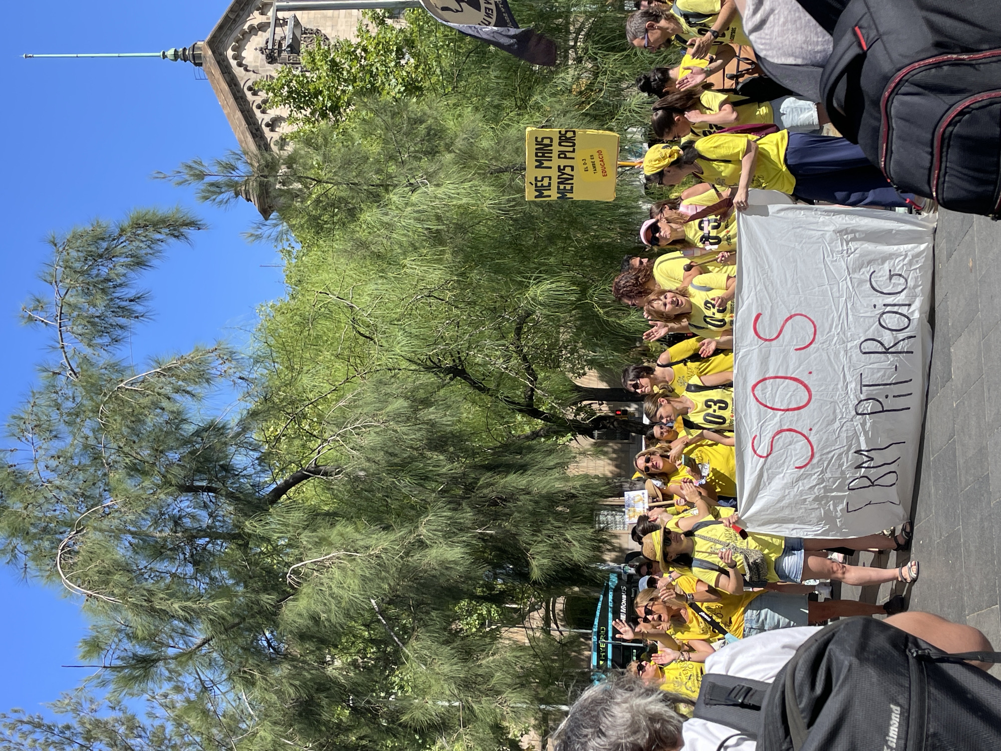In Barcelona, Spain, on a sunny day, a group of people are gathered, some holding a large white banner with red lettering that reads "S.O.S." and "TBM PIT. ROIG". Many individuals are wearing yellow shirts, some with "0-3" printed on them. One person holds a yellow sign with black text that says "MES MANS MENYS PLORS EL 0-3 TAMBE ÉS EDUCACIÓ". The background features lush green trees and a building with a spire. Some people are raising their hands, possibly in applause or protest. The scene suggests a public gathering or demonstration related to education, possibly at a school or educational institution given the "0-3" and "EDUCACIÓ" inscriptions.