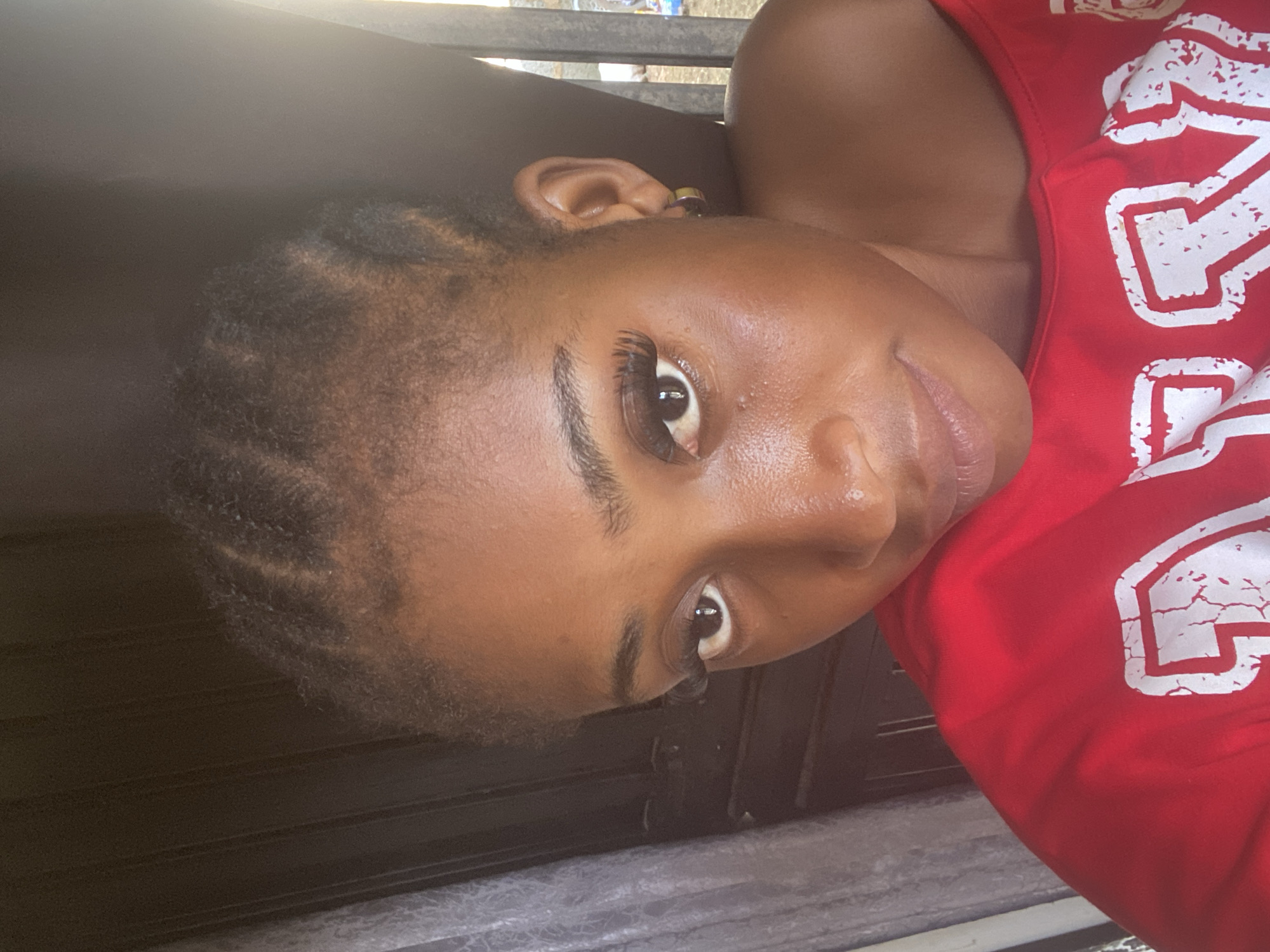 The image is a close-up selfie of a young woman with dark skin and braided hair. She is wearing a red off-the-shoulder top with white lettering. Her eyes are dark, and she is wearing false eyelashes. She has a small hoop earring in her left ear. The background is dark, with a sheer curtain on the left and what appears to be a metal fence or gate on the right. The lighting suggests it is daytime, possibly outdoors or near a window. The location in Abuja, Nigeria, is not explicitly depicted or evident from the image itself.