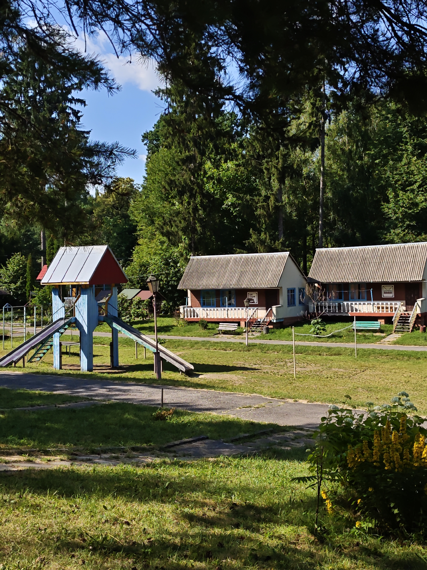 The image depicts a sunny day scene at a children's summer camp in Sosny, Ukraine. In the foreground, a blue and red playground structure with two slides stands on a grassy area. Two identical, single-story buildings with grey corrugated roofs and white-railed porches are visible in the mid-ground, set against a backdrop of dense green trees. A volleyball net is strung between two poles in front of the buildings. The ground is a mix of grass and paved pathways, with shadows indicating the presence of trees overhead. The overall impression is one of a peaceful, rustic outdoor recreational area.