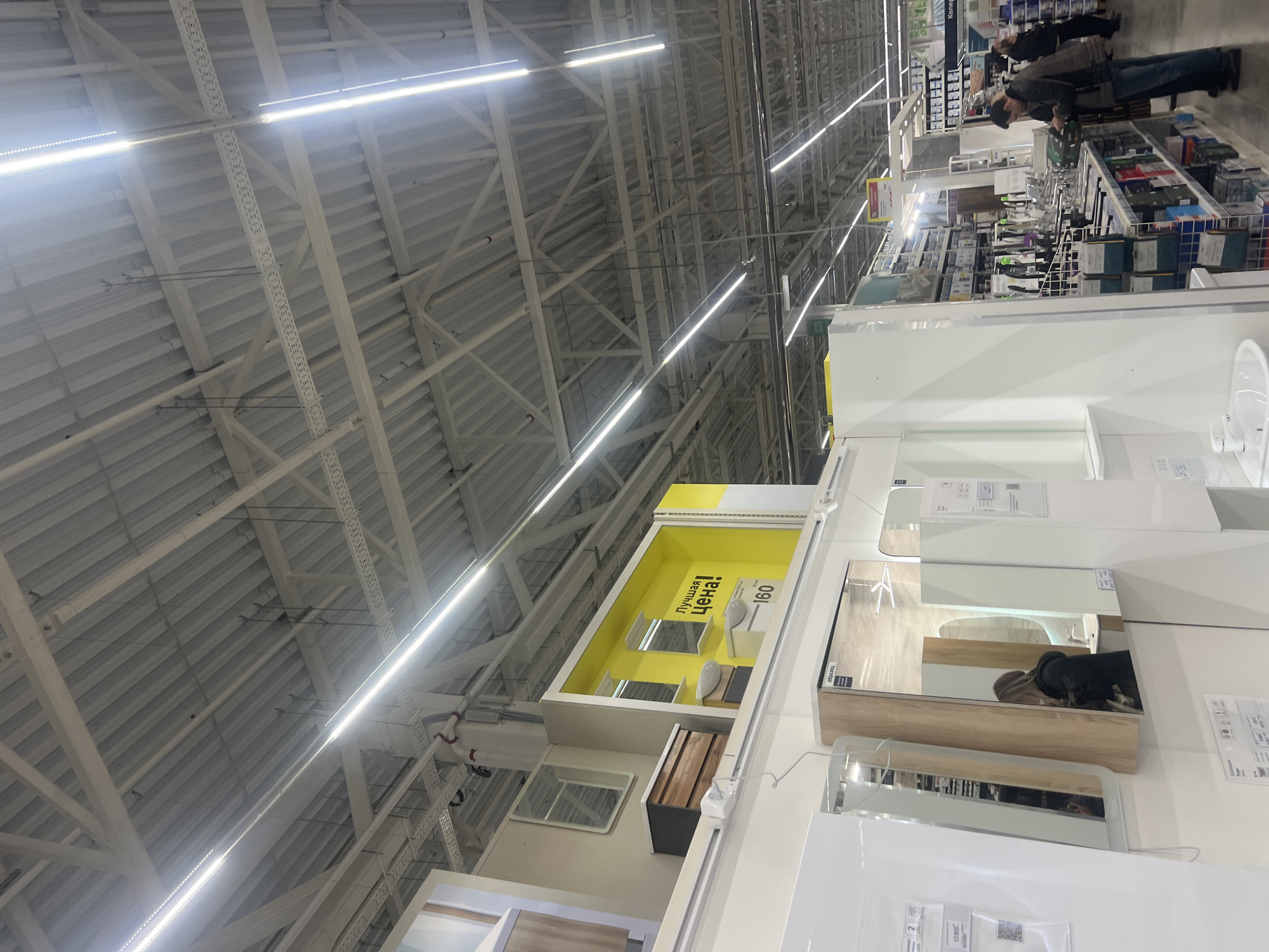 The image captures the interior of a store, likely a home improvement or furniture retailer in Izhevsk, Russia. The location is inferred from the presence of signage in Russian.
The primary scene focuses on a display area featuring bathroom fixtures, particularly mirrors and sinks. These are arranged against a white backdrop with product labels in Russian affixed to the surfaces. One display area, painted yellow, showcases mirrors and sinks along with the text "Лучшая Цена!", which translates to "Best Price!".
In the background, individuals are browsing other products. They are wearing casual clothing.
The store's architecture is visible, with a high, exposed ceiling structure. Fluorescent lighting fixtures illuminate the space.