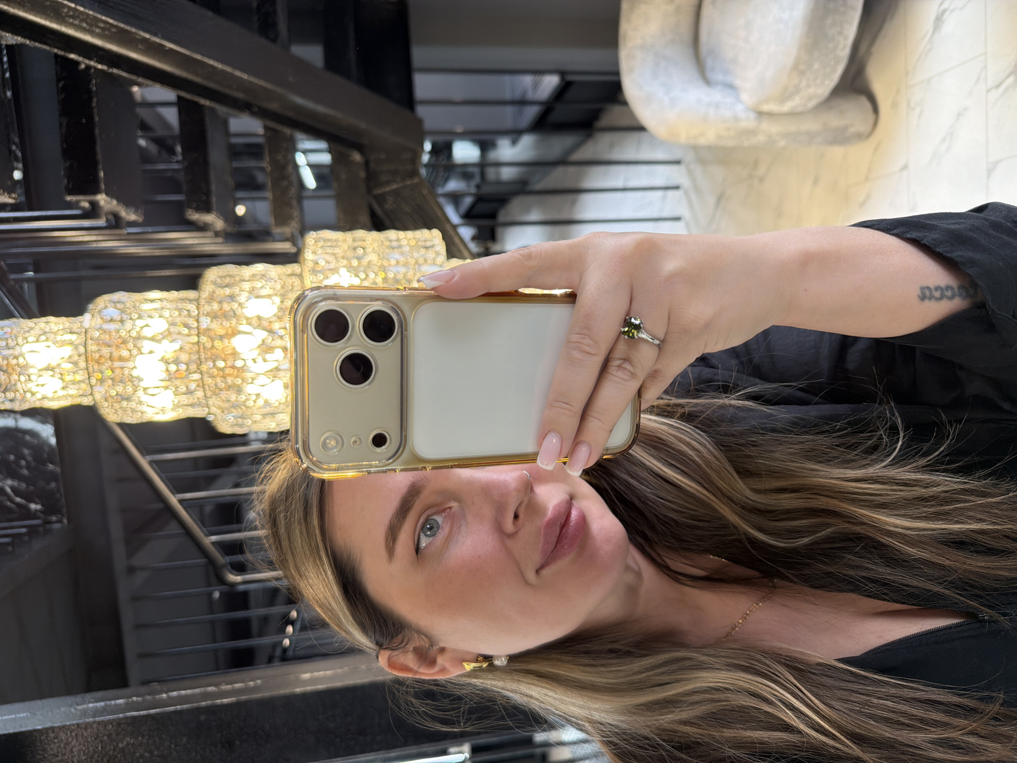 A woman is depicted taking a mirror selfie in an indoor setting, likely in Moscow, Russia, based on the provided context. The image is oriented vertically, showing the woman's face, right arm, and upper torso. She holds a gold-edged iPhone, equipped with a clear case and three camera lenses, in her right hand, positioned to capture her reflection.
Her face is angled slightly towards the camera, revealing light blue eyes, a subtle nose ring, and full lips. Her long, wavy hair, streaked with brown and lighter highlights, frames her face and extends over her shoulder. She wears a black top or jacket, and a delicate gold chain necklace is visible around her neck. A gold and pearl earring adorns her right ear. On the ring finger of her right hand, she wears a silver-toned ring set with a distinct green stone. Her nails are manicured with a French tip style.
A tattoo with an indistinct, stylized script is visible on the inner side of her right forearm.
The background, reflected in the mirror, features a large, ornate chandelier composed of numerous small, bright lights, suggesting a luxurious or upscale environment. Behind the chandelier, a dark metal staircase or railing structure is visible. To the upper right, a portion of an abstract, light-colored sculpture or furniture piece, possibly a white armchair, can be seen, along with a light-colored tiled floor. The overall impression is that of a well-lit, sophisticated interior space.