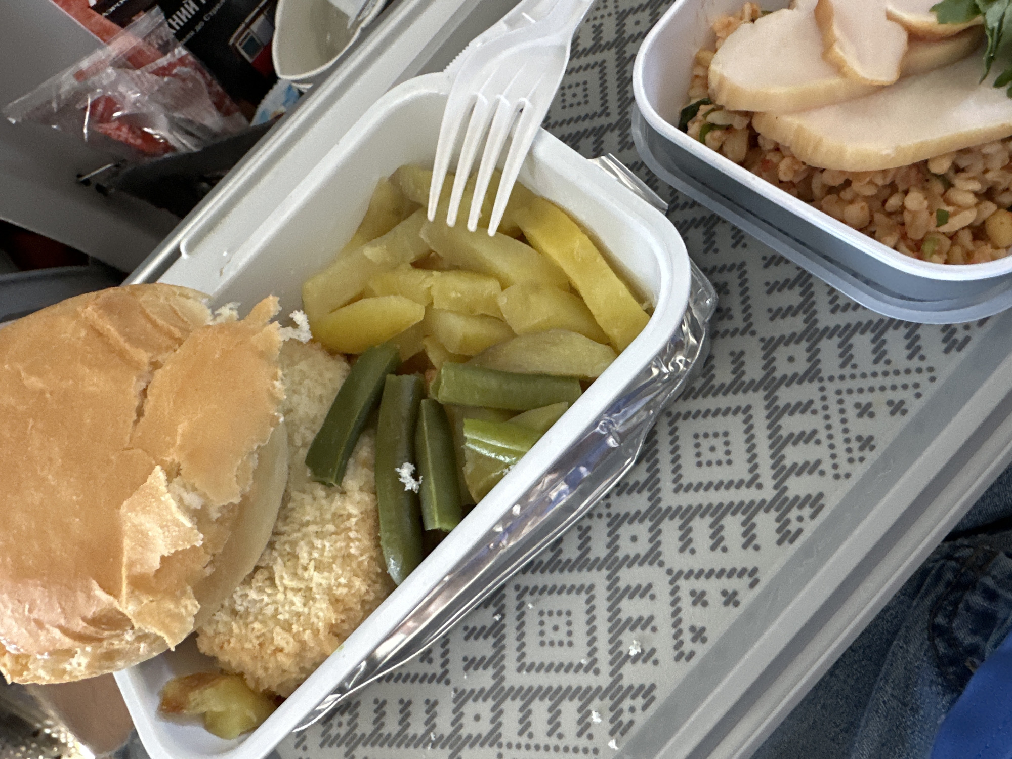 An overhead, close-up, static, eye-level shot captures an airline meal served on a tray. The meal consists of a bread roll atop a breaded chicken patty, accompanied by green beans and sliced potatoes. A white plastic fork rests on the potatoes. To the right, a second container holds rice with sliced chicken breast and a sprig of parsley. The tray is placed on a gray patterned surface, possibly a table or seat. The scene is indoors, suggesting a flight.