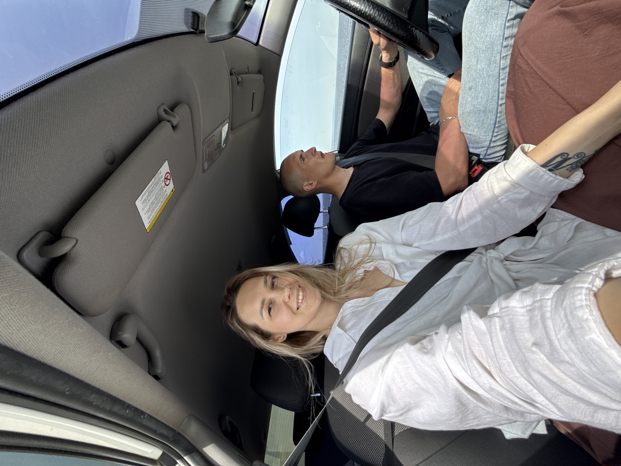 The image depicts a scene inside a car. A woman with blonde hair, wearing a white shirt and a seatbelt, is in the foreground, smiling towards the camera. Behind her and to the right is a man with a shaved head, wearing a black shirt and a seatbelt. His head is tilted back. The driver, partially visible, is holding the steering wheel. A part of their clothing can be seen. The interior of the car is visible, including the dashboard, sun visor, and side window. The location, Noviy, Russia, cannot be confirmed from the image.