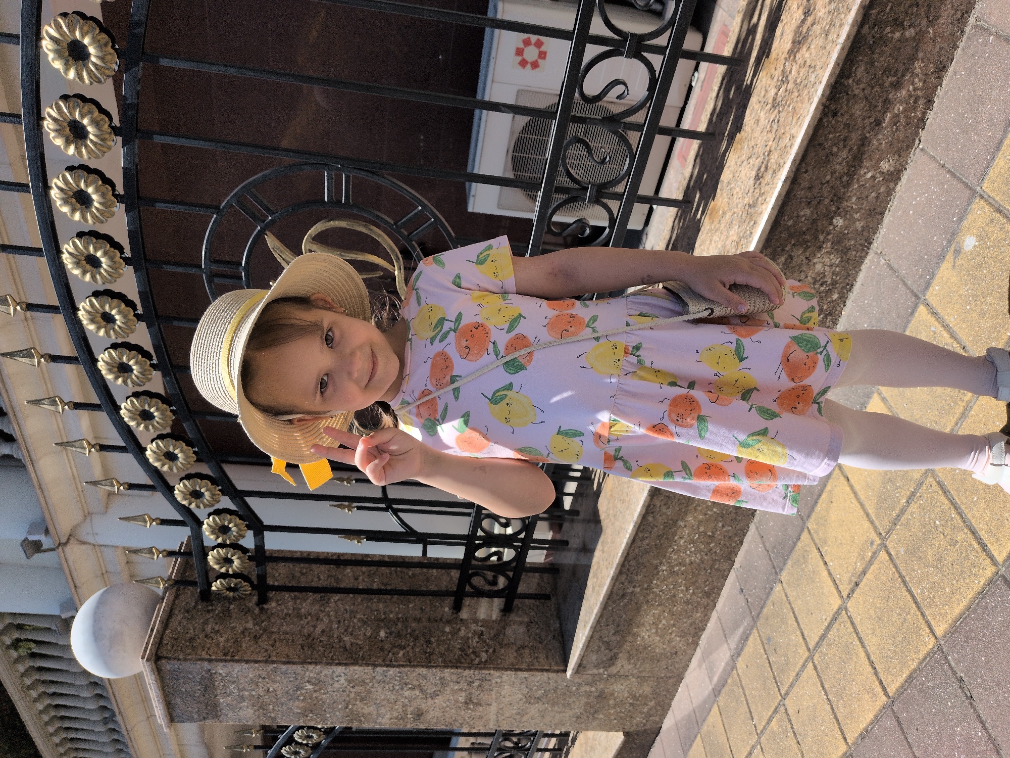 A young girl, approximately 4-6 years old, is captured in a full-length portrait, smiling directly at the camera on a bright, sunny day in Sochi, Russia. She wears a light-colored straw hat with a yellow band and a short-sleeved dress with a cheerful pattern of lemons and oranges on a pale pink or lavender background. Completing her outfit are white leggings and light-colored shoes, with a small woven handbag slung over her shoulder. Her right hand is raised, flashing a peace sign.
The girl stands outdoors next to an ornate black wrought-iron fence, which serves as a prominent background element. The fence is decorated with multiple gold-colored, flower-shaped medallions and pointed finials. Behind the fence, a dark brown building facade is visible, along with an air conditioning unit mounted on the wall. To the left, a spherical outdoor light fixture is attached to a stone or concrete pillar. The ground is paved with square tiles in alternating shades of brown and yellow-beige, and strong, defined shadows from the fence suggest it is midday or early afternoon with clear skies. The scene evokes a cheerful and summery atmosphere. No discernible text is visible in the image.