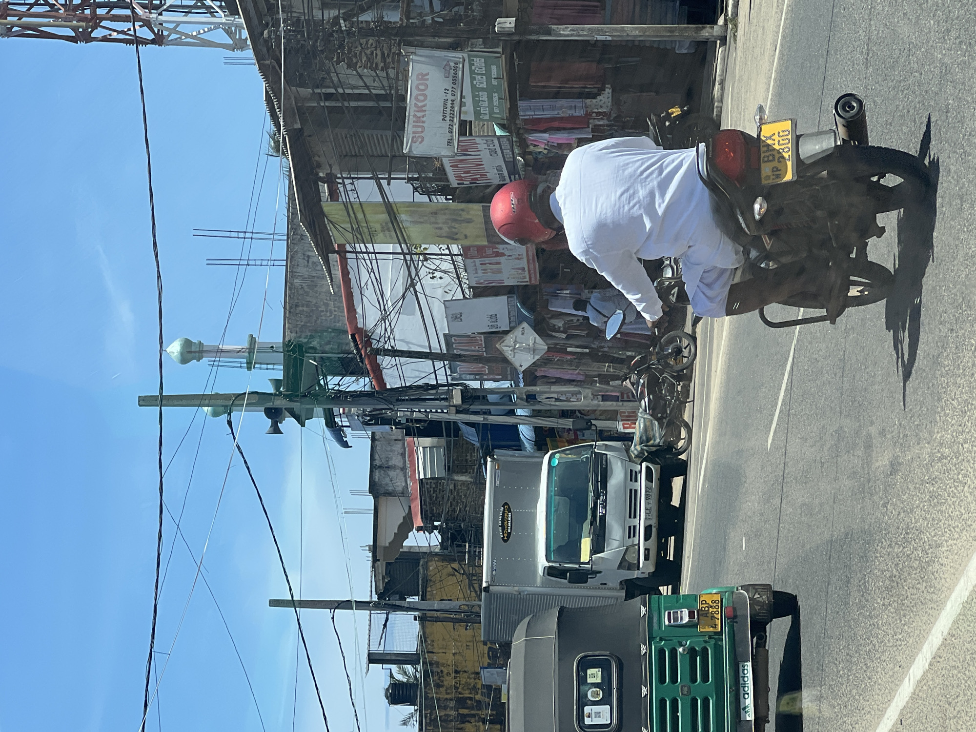 The image depicts a street scene in Pottuvil, Sri Lanka. A man wearing a white shirt and red helmet is riding a motorcycle with license plate "MP BHX 2800." A green three-wheeled vehicle ("tuk-tuk") with license plate "BP 7888" and an Adidas logo on the mud flap is visible. A white truck with "TRAFFIC RENTALS" writing is also in the scene. Overhead power lines and signage including "SUKKOOR, POTTUVIL-12" can be seen. There is a mosque minaret in the background.
