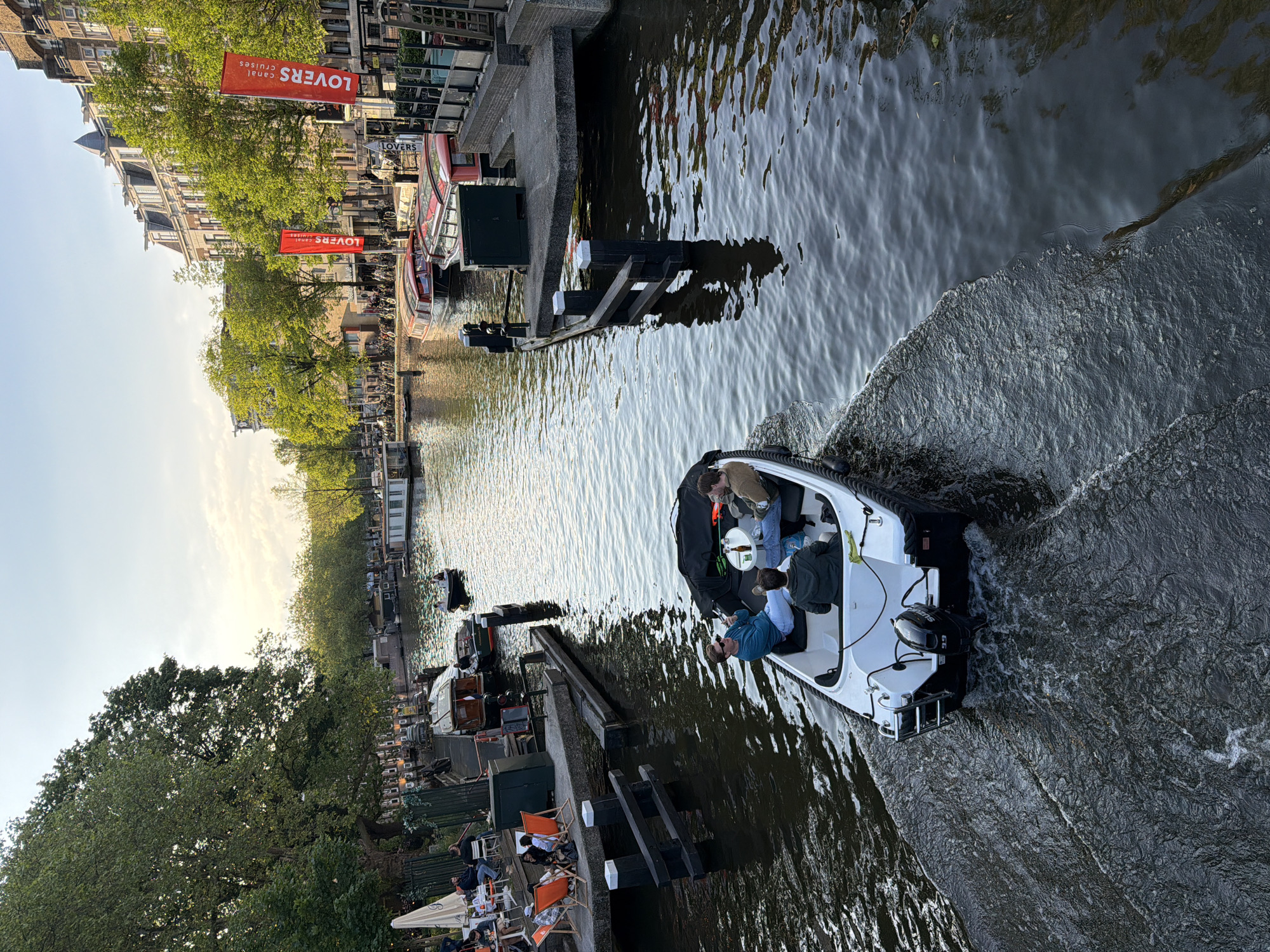 The image shows a canal in Amsterdam, Netherlands, with a boat moving through the water. The boat has a white hull and is occupied by at least two people. One person is wearing a blue shirt, and there is a small table inside the boat with what appears to be a bottle on it.
Along the canal's edge, there are buildings, trees, and a waterfront area where people are sitting in chairs at tables, some under an umbrella. There are other boats docked or moving in the canal, and signs advertising "Lovers Canal Cruises" are visible. The water is dark, with visible ripples and waves created by the movement of the boat in the foreground. The sky is clear and suggests it is either early morning or late afternoon.