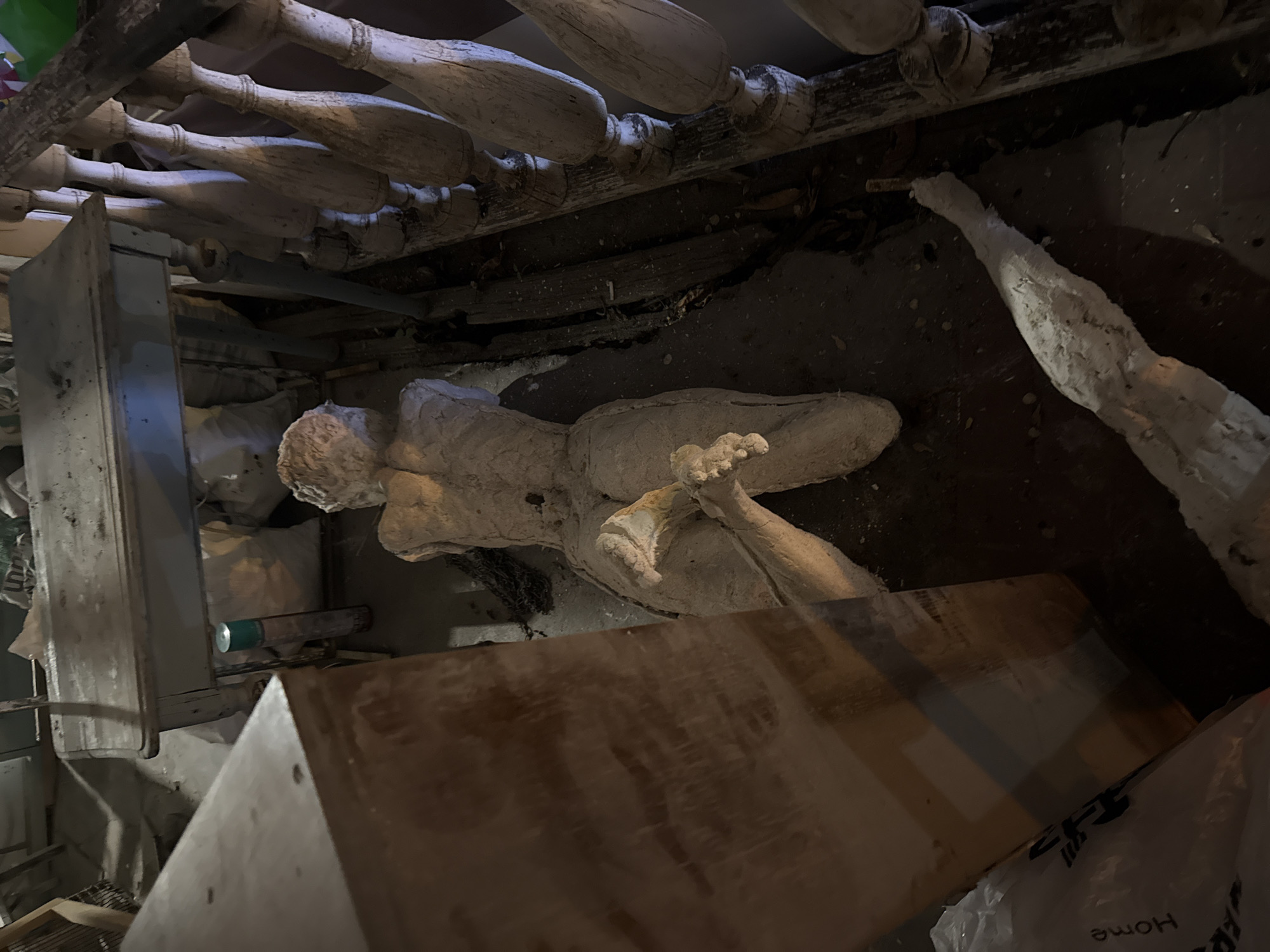 The image depicts a seemingly cluttered, confined space. At the center of the view lies a deteriorated plaster cast of a human figure, possibly a sculpture or study model, lying horizontally. An unidentified cylindrical container with a green cap is visible. Various pieces of what appear to be furniture components, including wooden spindles and panels, are scattered around the plaster figure. The word "Home" is visible on a white plastic bag in the bottom right corner.
The image was taken in Yerevan, Armenia.