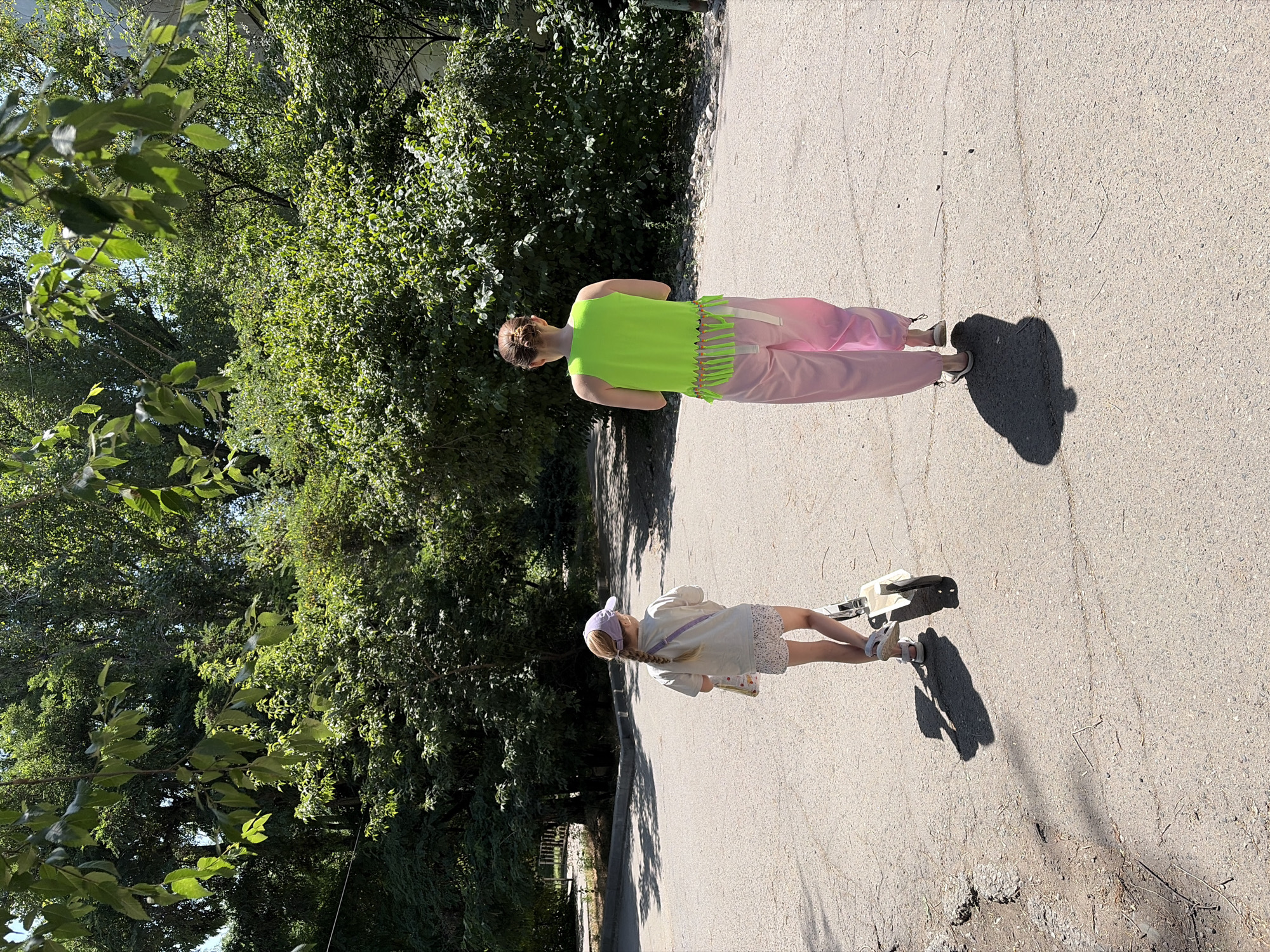 In an outdoor urban setting, two individuals are viewed from behind, walking away from the camera. The individual on the left is a child wearing a light-colored t-shirt, patterned shorts, and a purple baseball cap. They are also holding onto a white scooter. The individual on the right is an adult wearing a bright green sleeveless top with fringe detailing and loose pink pants. They are walking on a paved surface, possibly a path or sidewalk, with lush green trees and foliage surrounding the area, suggesting a park or a tree-lined street. The lighting indicates it is daytime, with shadows cast on the ground.