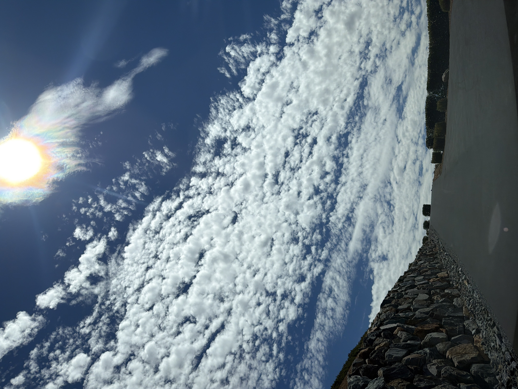 This image depicts a clear blue sky filled with scattered white, cumulus clouds. The sun is visible at the top center of the frame, partially obscured by a wispy cloud that displays iridescence, creating a sun halo effect. In the foreground, a paved road curves upwards, flanked on the left by a stone retaining wall constructed of large, irregular grey rocks. The wall is topped with sparse vegetation. On the right side of the road, a well-maintained hedge forms a border, with smaller bushes spaced along it. The overall scene suggests a road leading into a developed area, possibly a residential or recreational property, under a bright, sunny sky. The location is specified as El Dorado Hills, United States.