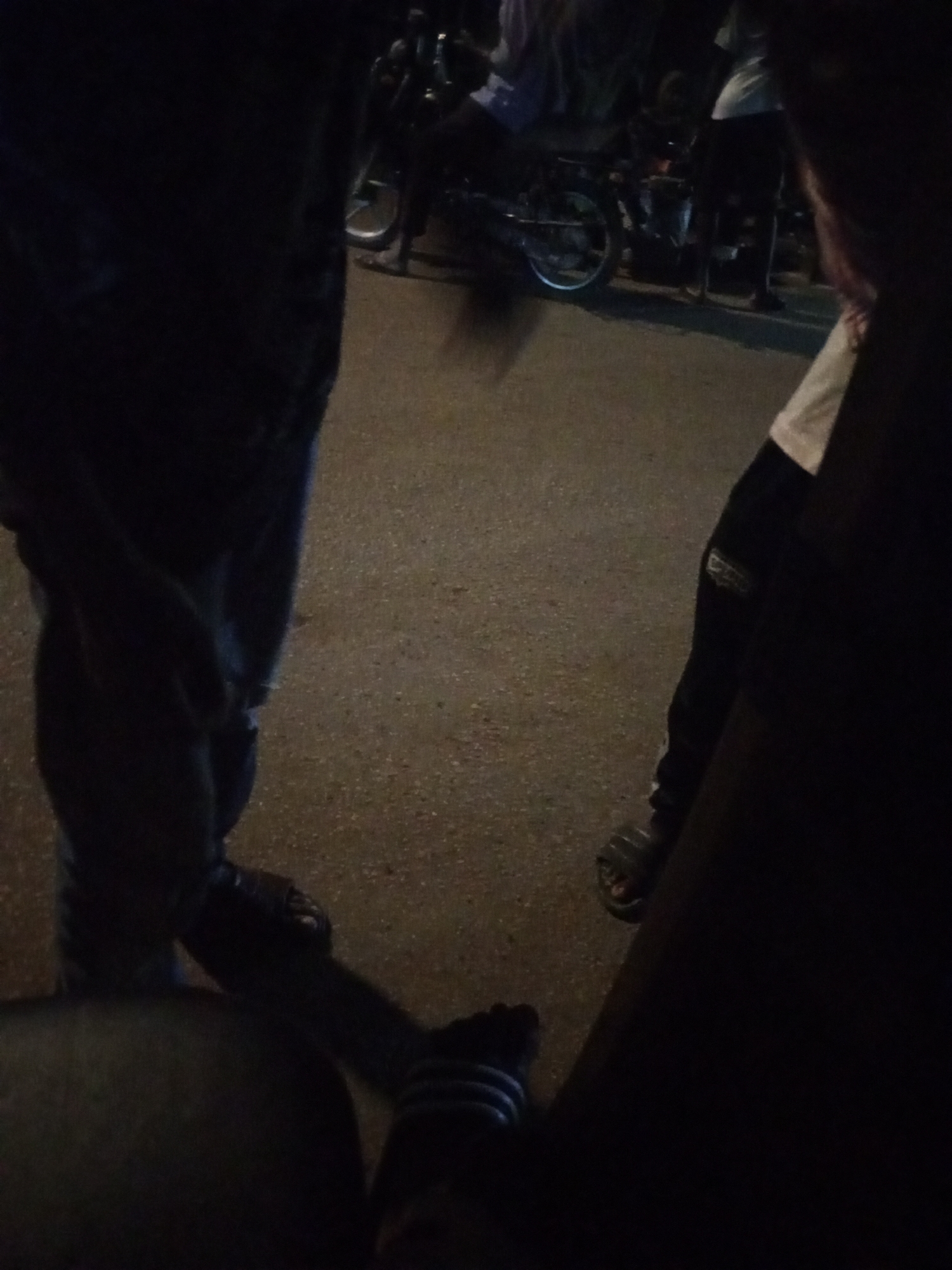 The photograph presents a dark, nighttime outdoor scene, likely on an asphalt or paved street. In the immediate foreground on the left, the lower half of a person is visible, wearing dark trousers or jeans and dark sandals or slippers. On the right side, another individual is partially seen, mostly their right leg and torso, also dressed in dark attire with sandals. A small white patch, possibly a logo or design, is discernible on their upper right arm.
In the mid-ground and background, several motorcycles or mopeds are dimly visible, appearing to be stationary or parked. Their wheels and frames are faintly outlined against the low light. A few other indistinct human figures can be observed standing or sitting near these vehicles, suggesting a casual gathering or street activity.
The extreme darkness indicates the time of day is night or very late evening. No specific weather conditions can be determined. The scene appears to be a street or public area, consistent with an urban or semi-urban setting like Maiburiji, Nigeria. No discernible emotions are conveyed as faces are not visible, and there is no readable text anywhere in the image.