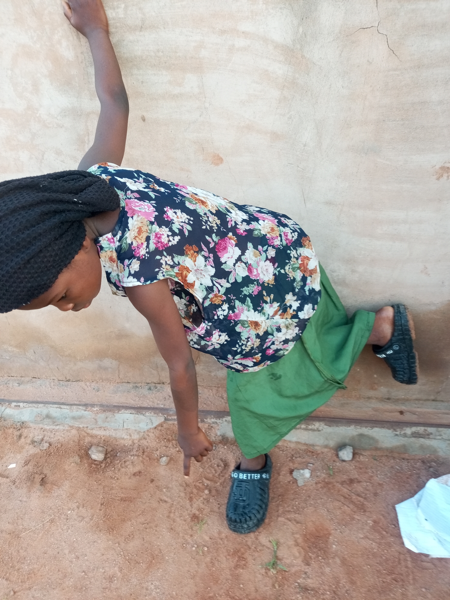 A young person with dark skin, wearing a floral print shirt and a green skirt, leans against a textured, light-colored wall. Their right arm is extended upwards, resting on the wall, while their left arm is bent, and their left hand is pointing downwards at the sandy ground. The person has braided dark hair and is wearing black clogs on their feet. The environment appears to be outdoors, with a dusty, reddish-brown ground scattered with small stones and a white piece of cloth in the lower right corner. The wall has visible cracks and imperfections. The scene is set in Tempure, Nigeria.