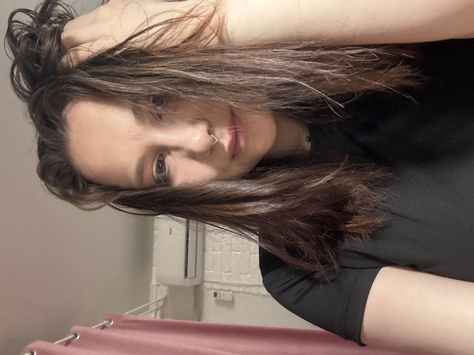 An adult female is depicted in a close-up, frontal self-portrait taken indoors. She has dark brown, slightly disheveled hair, with strands partially obscuring her face. Her left hand is raised, fingers entangled in her hair on the top-right side of her head. She wears a black short-sleeved t-shirt and features a silver-toned, V-shaped septum piercing. Pink eyeshadow is visible on her right eyelid. The background to her left consists of a white painted brick wall with a white Haier-branded split-system air conditioning unit mounted above it. A section of light pink curtain with metal grommets is visible on the far left. The specific location within the city of Saratov, Russia, is not discernible from the provided image.