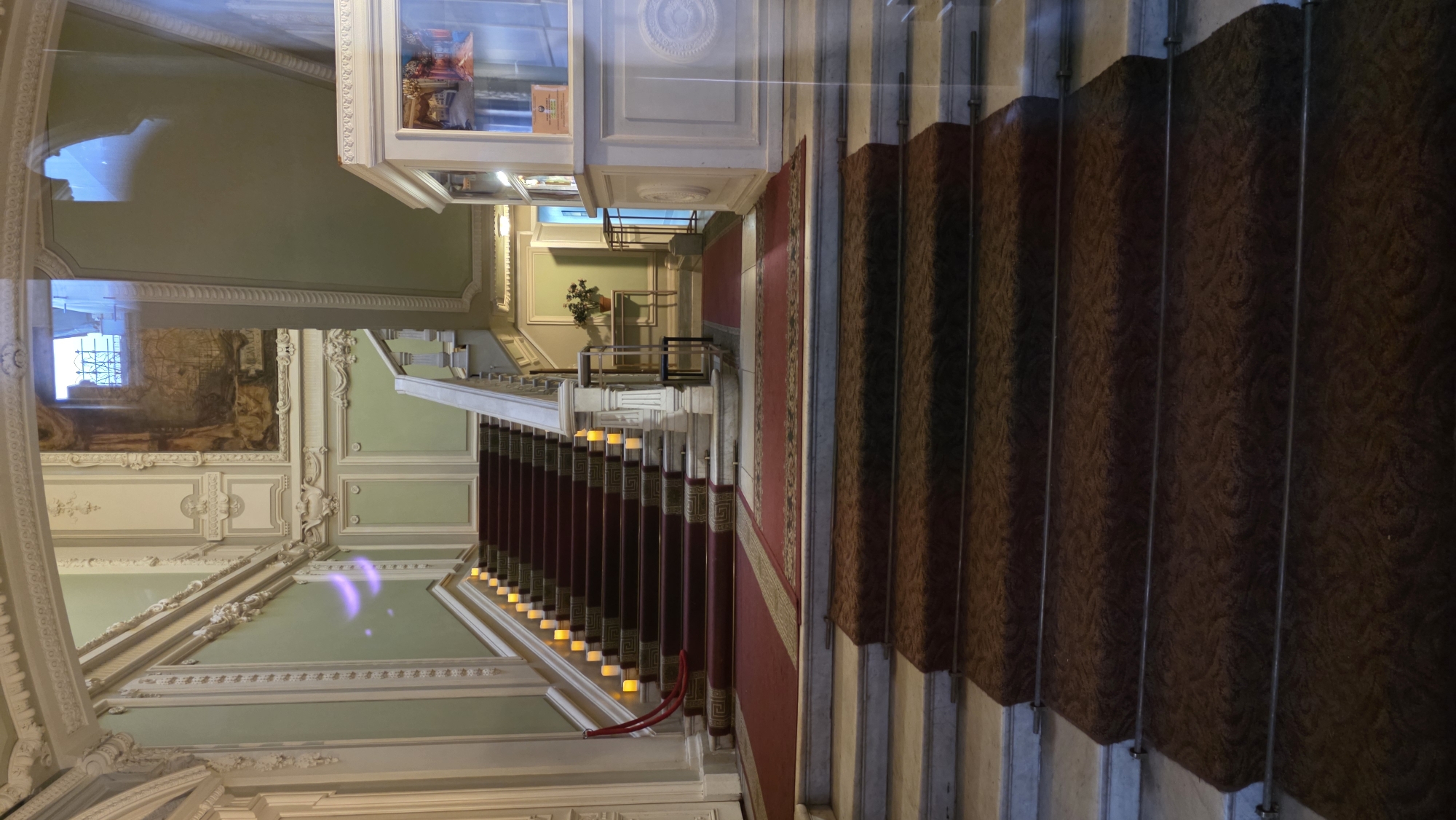 The image shows a staircase in a grand interior, likely located in Saint Petersburg, Russia. The staircase is carpeted with a red runner that has ornate designs and is flanked by white marble or stone steps. A section of the staircase has a brown carpet. The interior is decorated with intricate moldings, panels, and ornamental details on the walls and ceiling. There are murals on the walls. There is a cabinet on display. Overall, the scene is luxurious and reflects a historical setting. The image is vertically oriented, giving an upward view of the staircase.