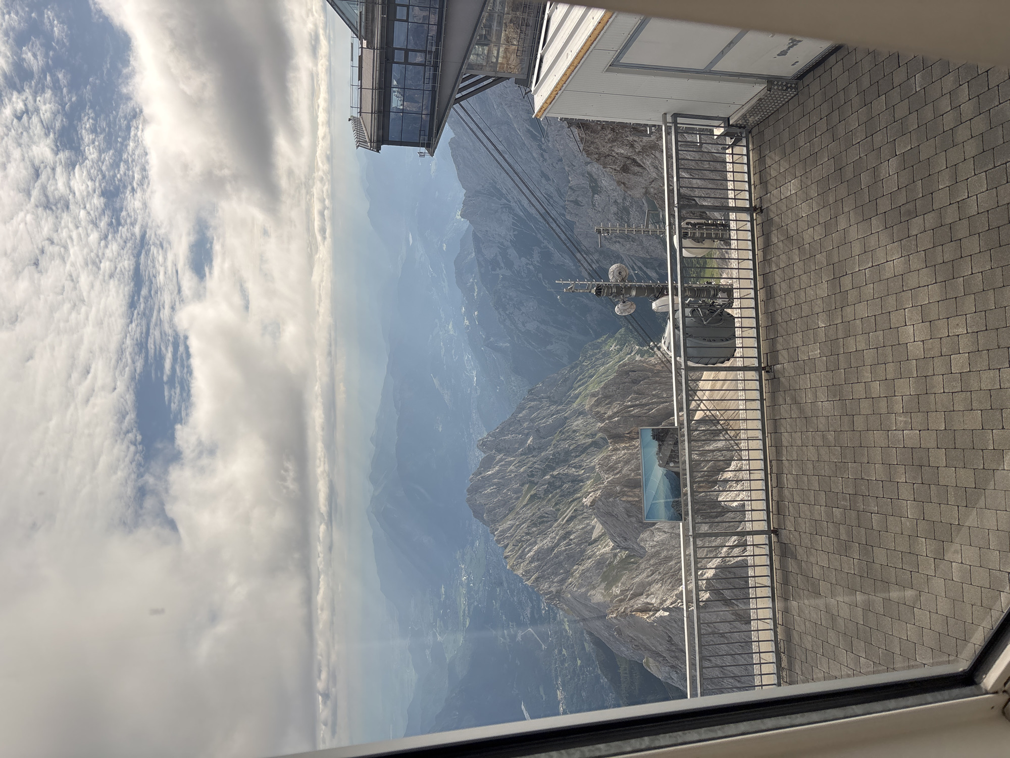 This is a photograph taken from inside a building, looking out through a window at a mountainous landscape. In the foreground, there is a paved patio area with a metal railing. Beyond the railing, a steep, rocky mountain peak dominates the view. In the distance, more mountains recede into a hazy blue atmosphere, with a valley and scattered buildings visible. The sky is partly cloudy with sunlight breaking through. To the right of the frame, a modern building with large glass windows is visible, cantilevered over the mountainside, and a white building with corrugated siding is also present. There are also some communication towers and cables in the midground. The scene appears to be from a high-altitude observation point or a mountain resort. The view is consistent with the Zugspitze, Germany's highest peak, which is located near Ehrwald, Austria, and accessible by cable car.
