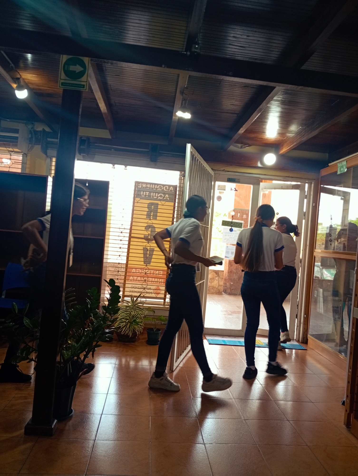 The image depicts an interior scene, possibly a commercial establishment, with a warm, tiled floor and a wooden ceiling. Three young individuals, appearing to be students or employees, are the main subjects. One person, viewed from the side, wears glasses and a white shirt with blue accents, holding a book or tablet. Another individual, facing away and with long hair, also wears a white shirt with blue accents and dark pants, walking through an open doorway. A third person, partially visible, wears a similar uniform and walks behind the second individual.
The environment includes indoor plants in pots, suggesting a well-maintained space. A sign with an arrow pointing right is visible on a pillar, likely indicating direction. Through the open doorways, a glimpse of the exterior suggests a street or pathway with a vehicle. The lighting suggests it is daytime, with natural light entering through the doors and artificial lighting from the ceiling. The overall atmosphere is casual and active, with the individuals engaged in movement. The location is identified as Juan Griego, Venezuela.